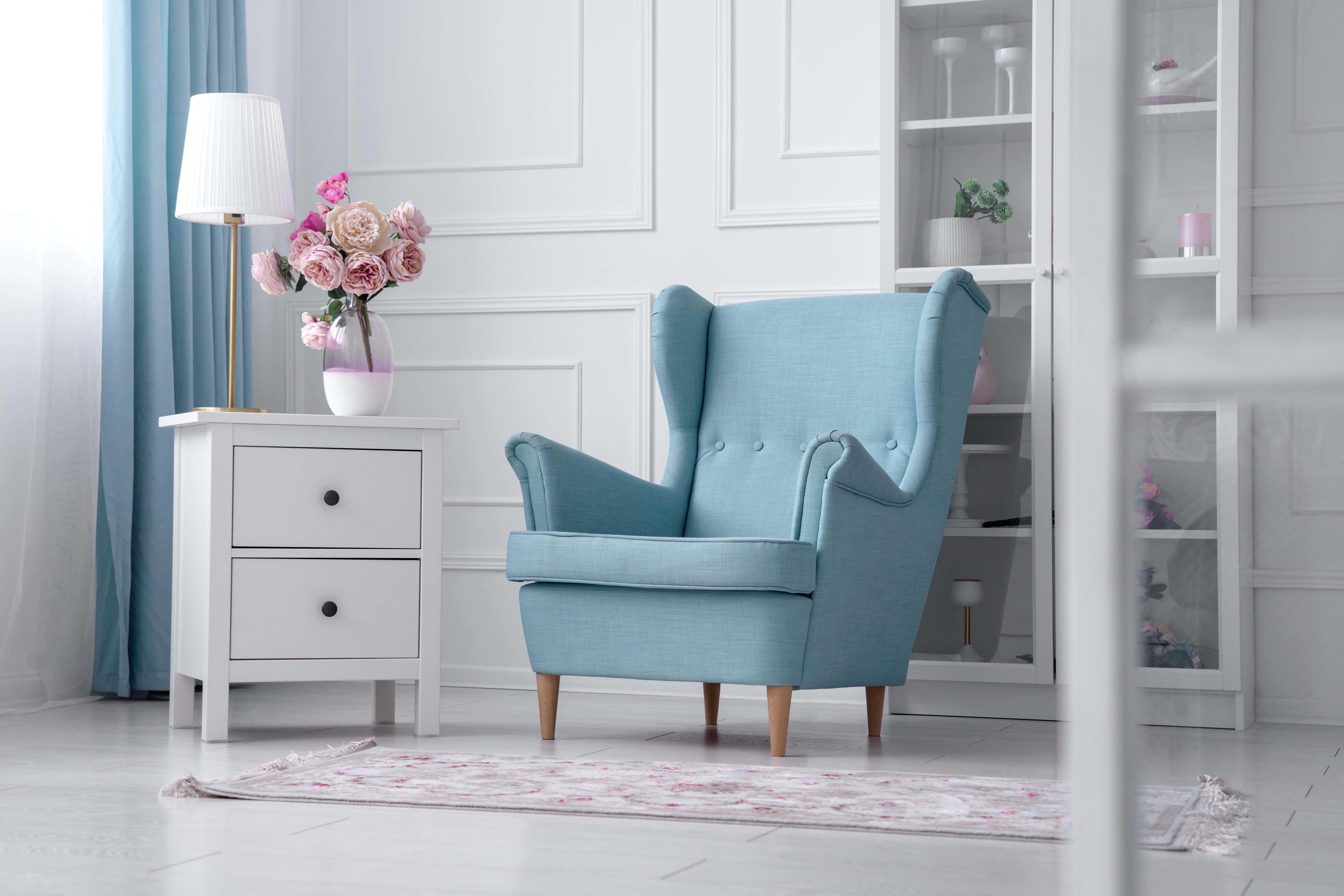 A sapphire-blue wingback armchair angled beside a mirrored dresser with fresh flowers, in a bright, classically trimmed room.