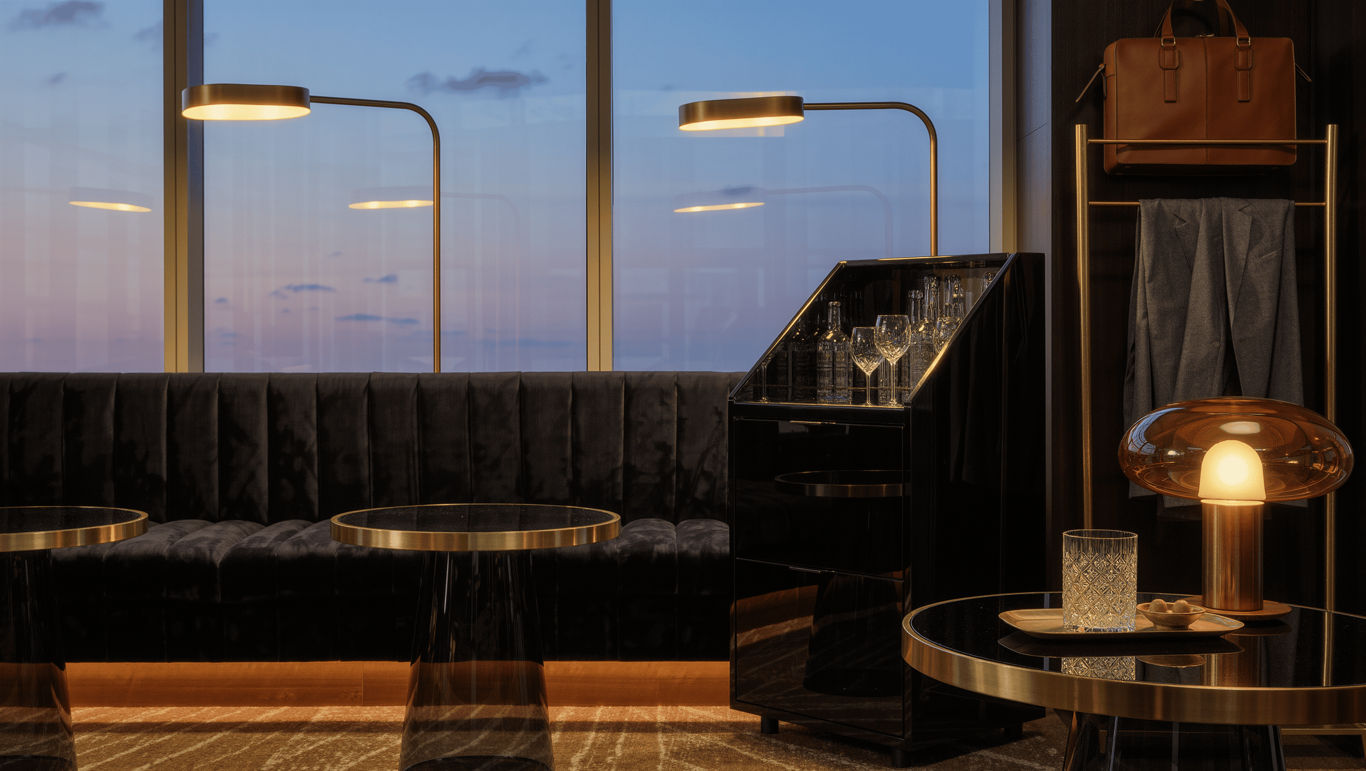 A hospitality-style lounge with a charcoal velvet banquette, black-lacquer drinks cabinet, brass rail with a leather bag, and warm lamps at dusk.