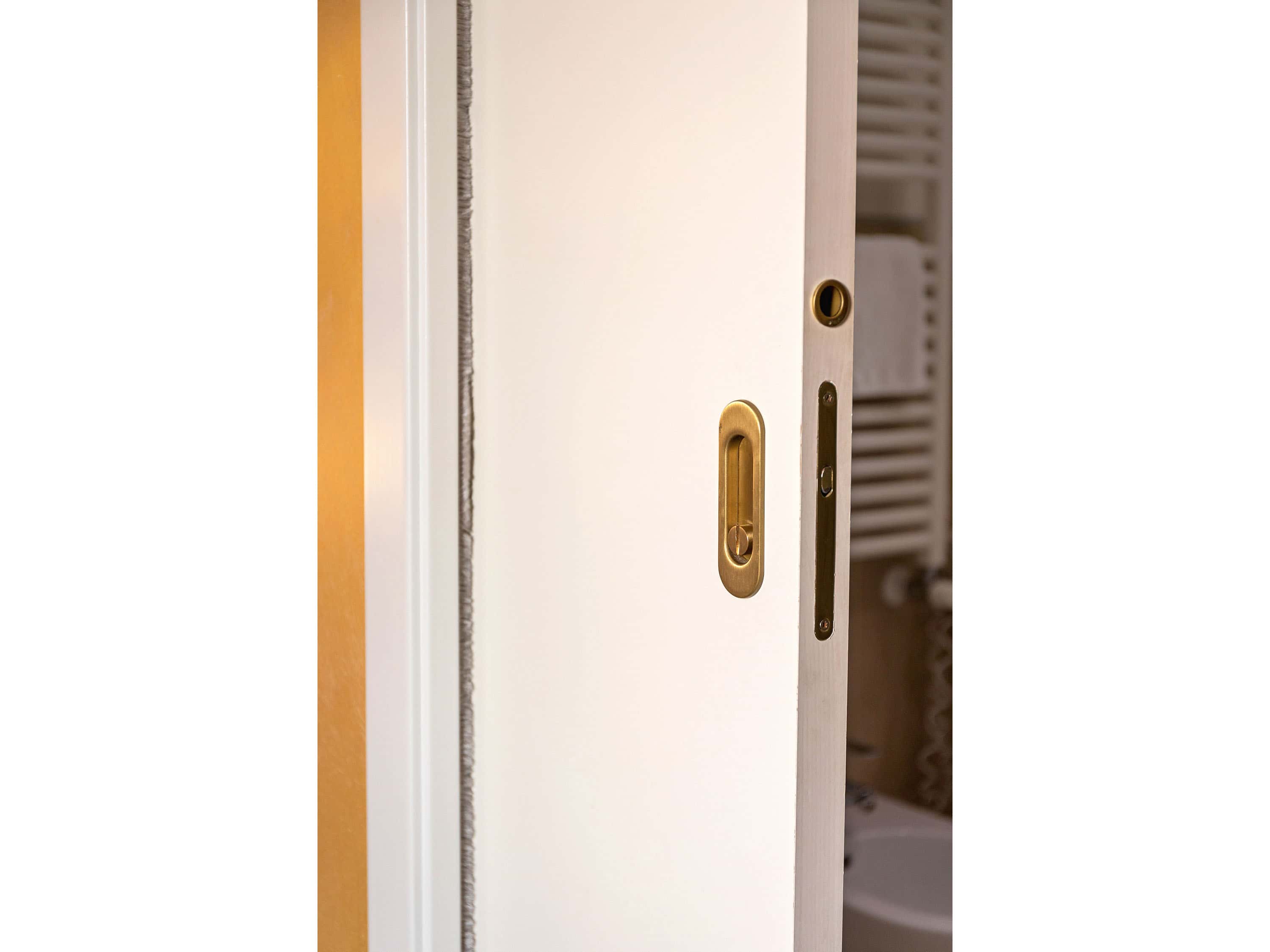 A white pocket door fitted with a polished brass oval pull and thumb-turn lock, slid partway into the wall pocket with a tiled bathroom visible beyond.