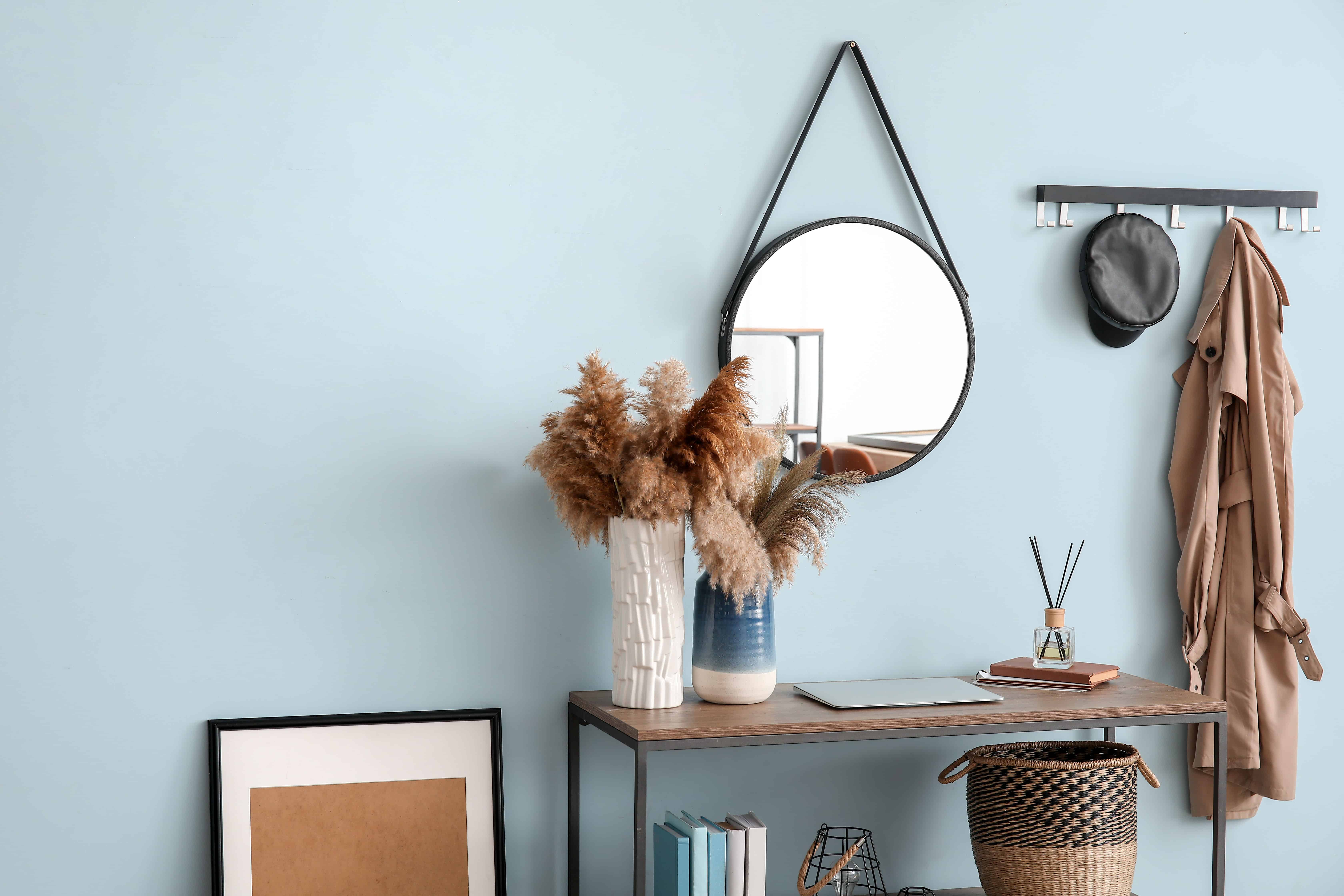A pale blue entryway with a round mirror above a metal console table holding a vase of pampas grass and a woven basket below.