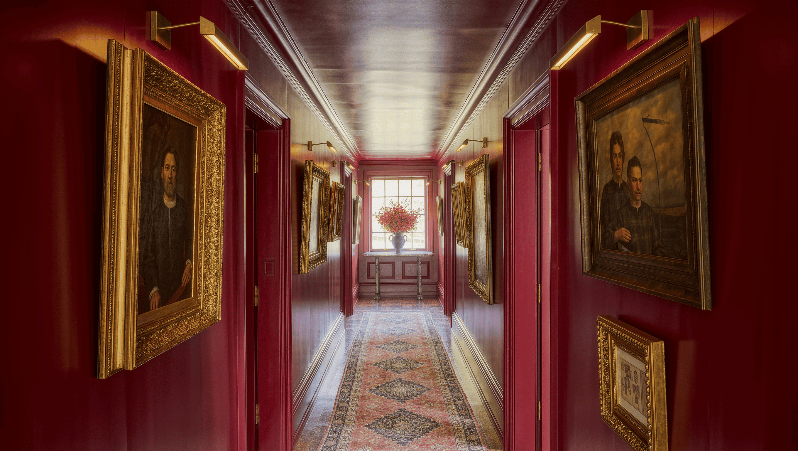 Long oxblood hallway hung salon-style with gilt-framed art under brass picture lights, a runner rug leading to a bright window.