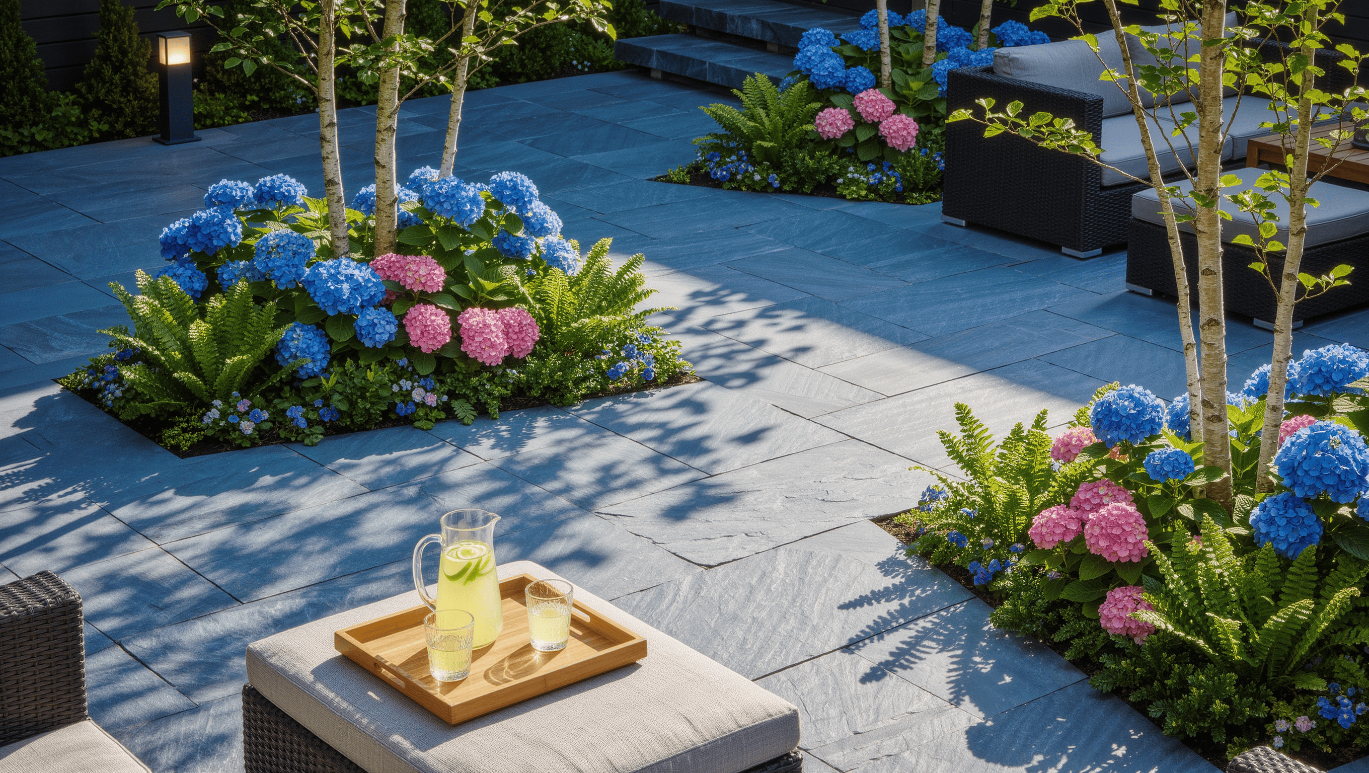 A large bluestone patio with two planted islands of hydrangea, ferns, and birch breaking up the hardscape, with a seating group in midday sun.