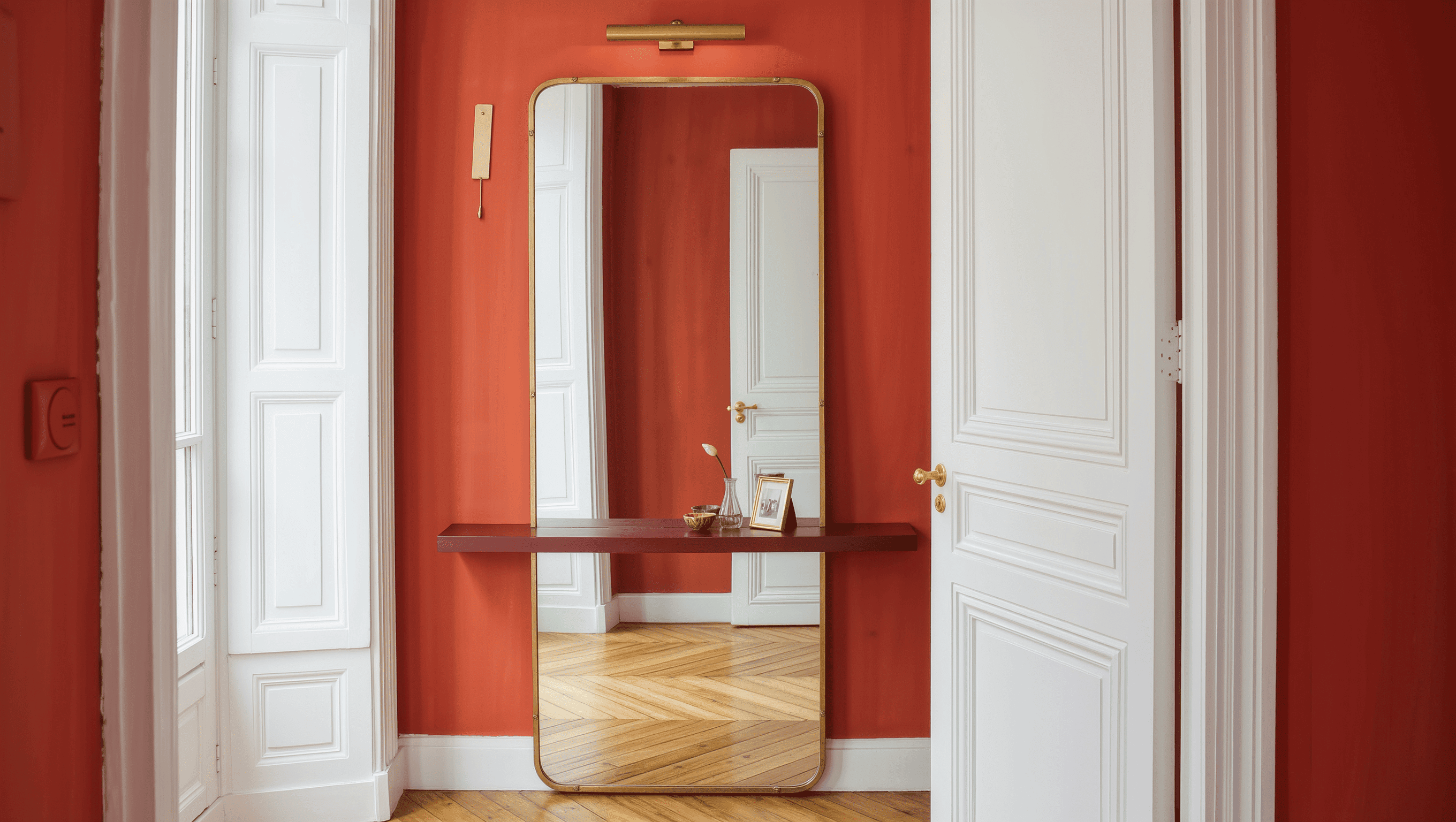 A narrow wall between two paneled doors holding a tall brass mirror, a shallow oxblood ledge, and a small key dish.