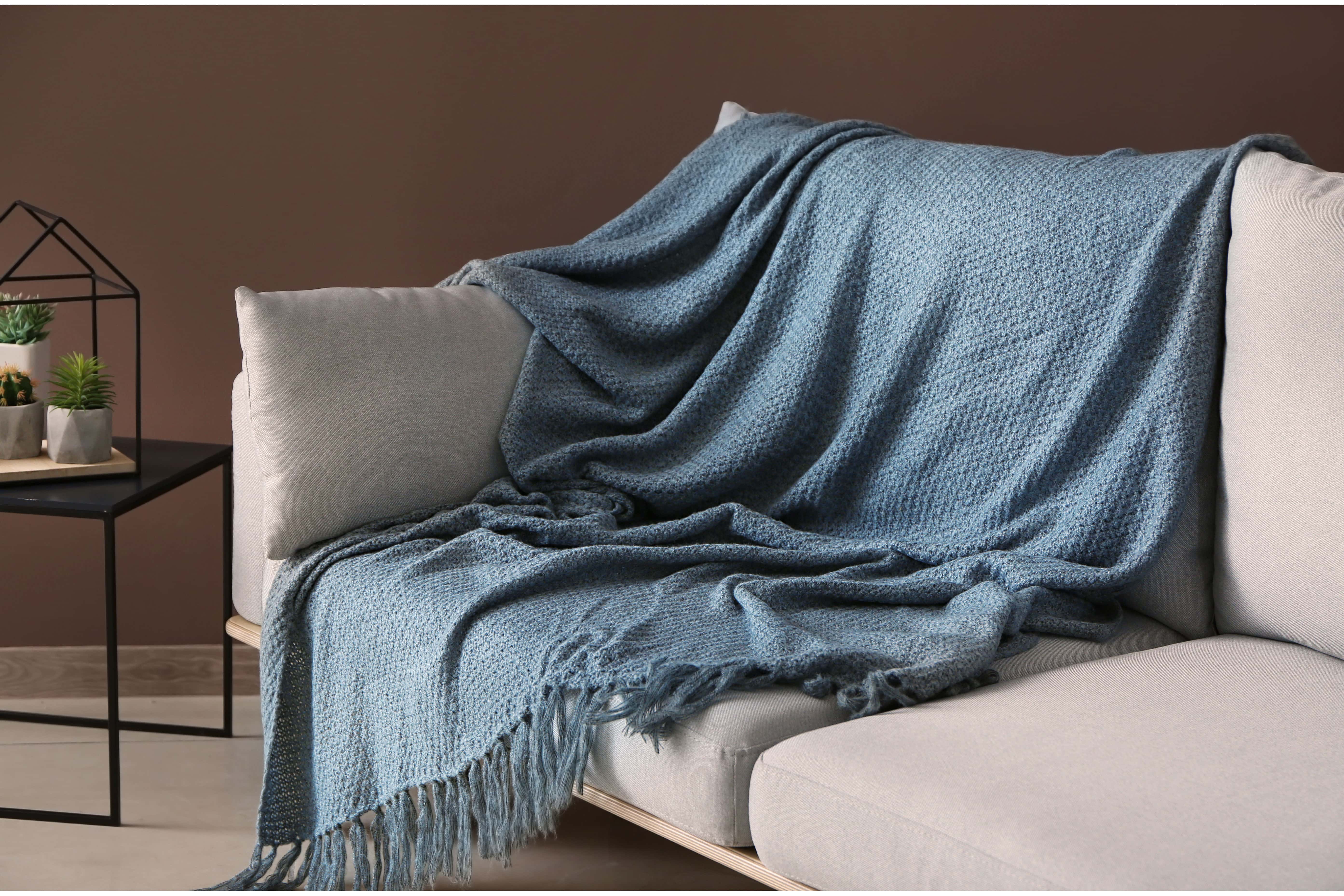 A chunky woven throw with a fringed edge draped over the arm of a linen sofa in a warm, softly lit room.