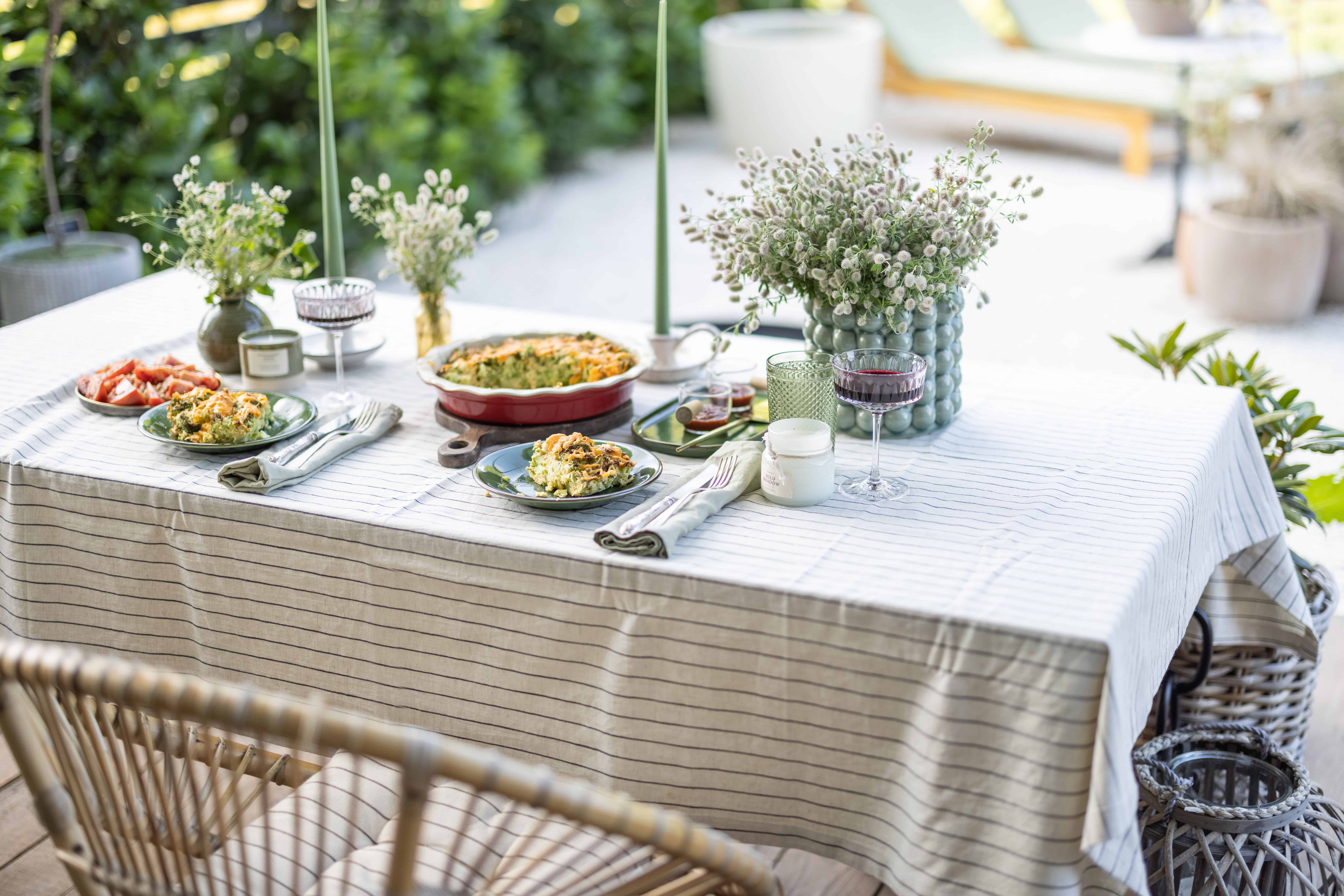 A linen-covered garden table set with plates of food, glassware, and a jug of white wildflowers, flanked by woven rattan chairs amid greenery.