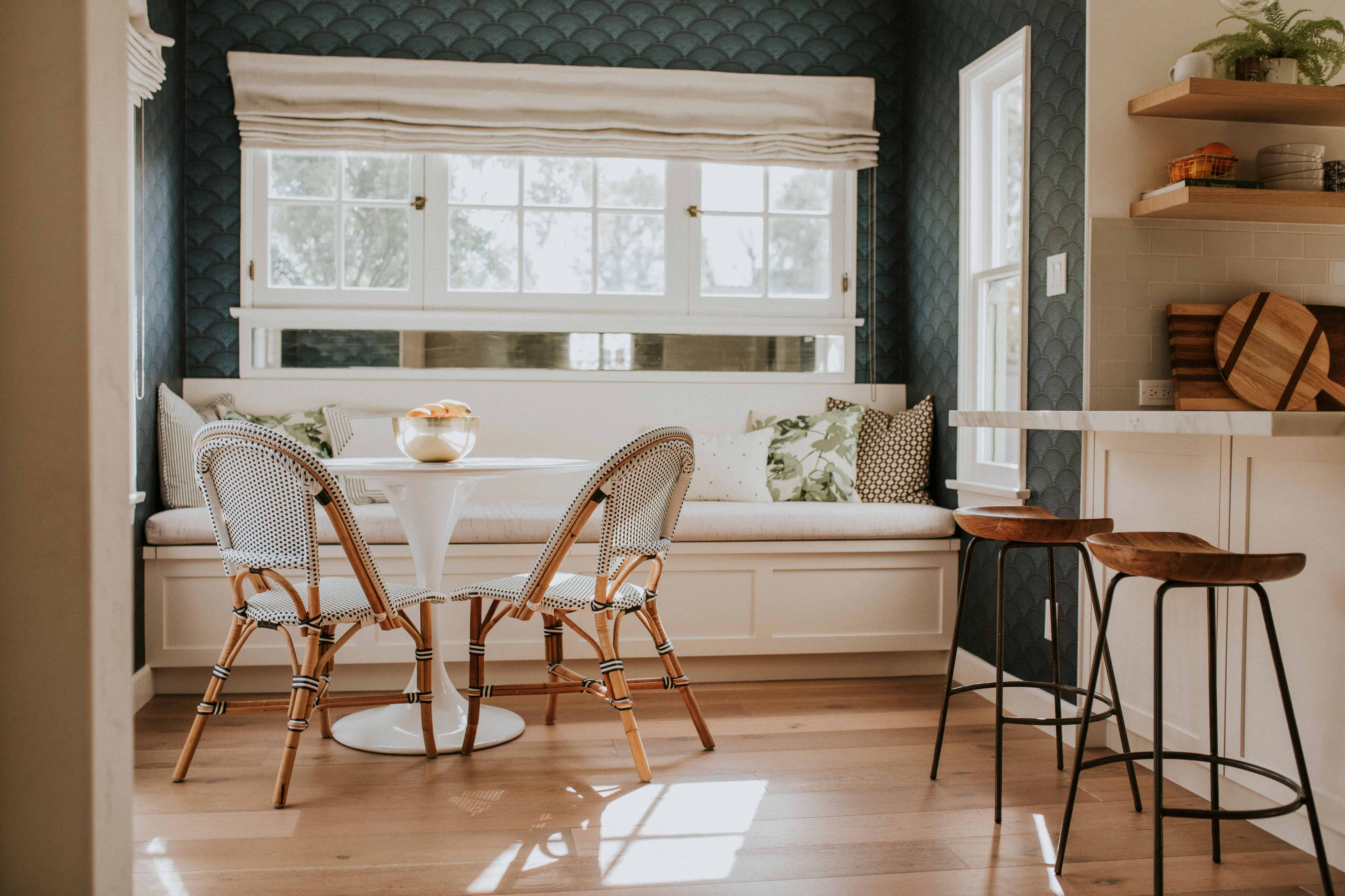 A built-in white banquette with striped pillows sits beneath a bay window against a richly patterned navy wallpaper, ringed by rattan chairs at a round marble-top table.