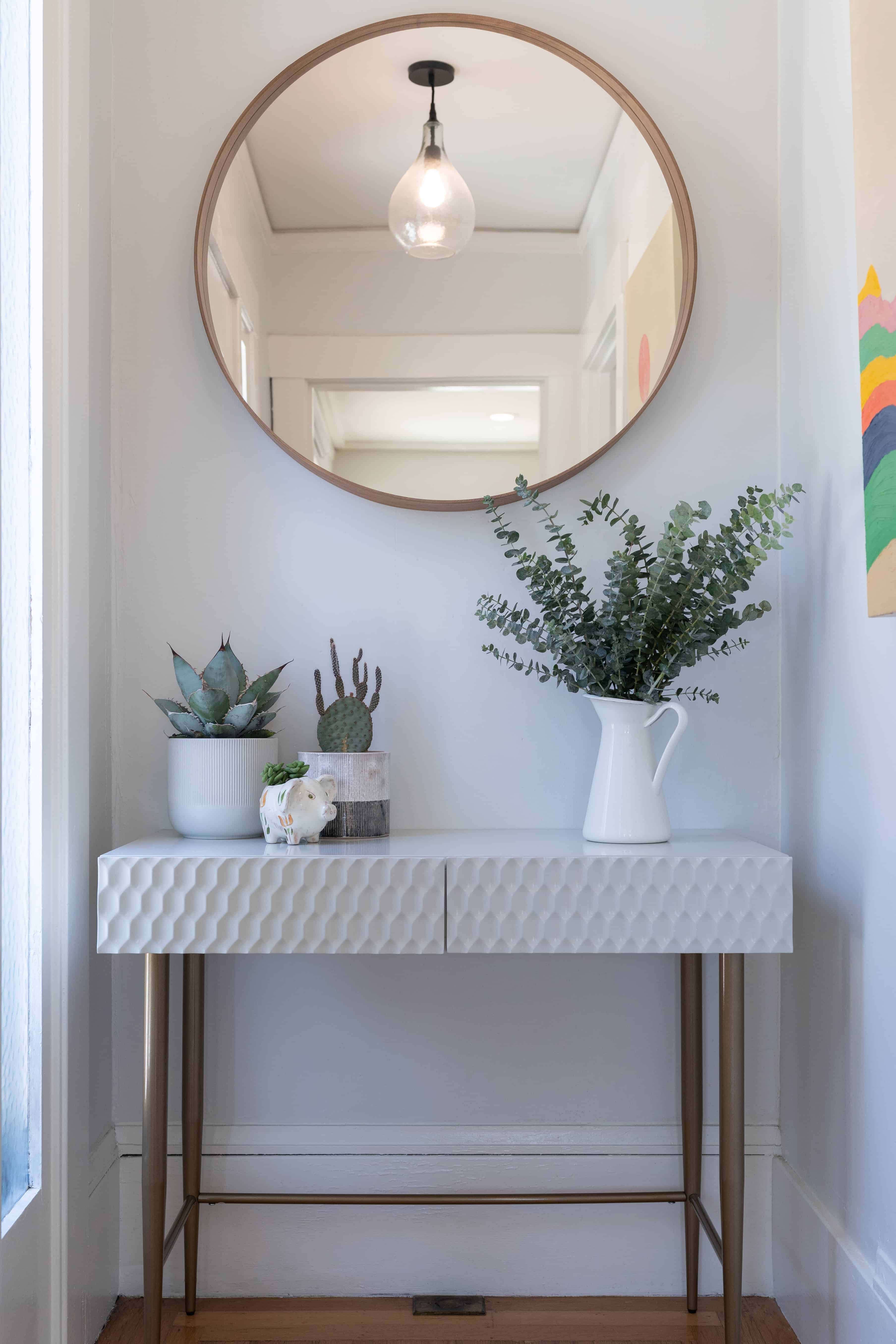 A fluted marble-look console table sits below a round brass-framed mirror, styled with a stoneware vase of eucalyptus and small potted succulents.