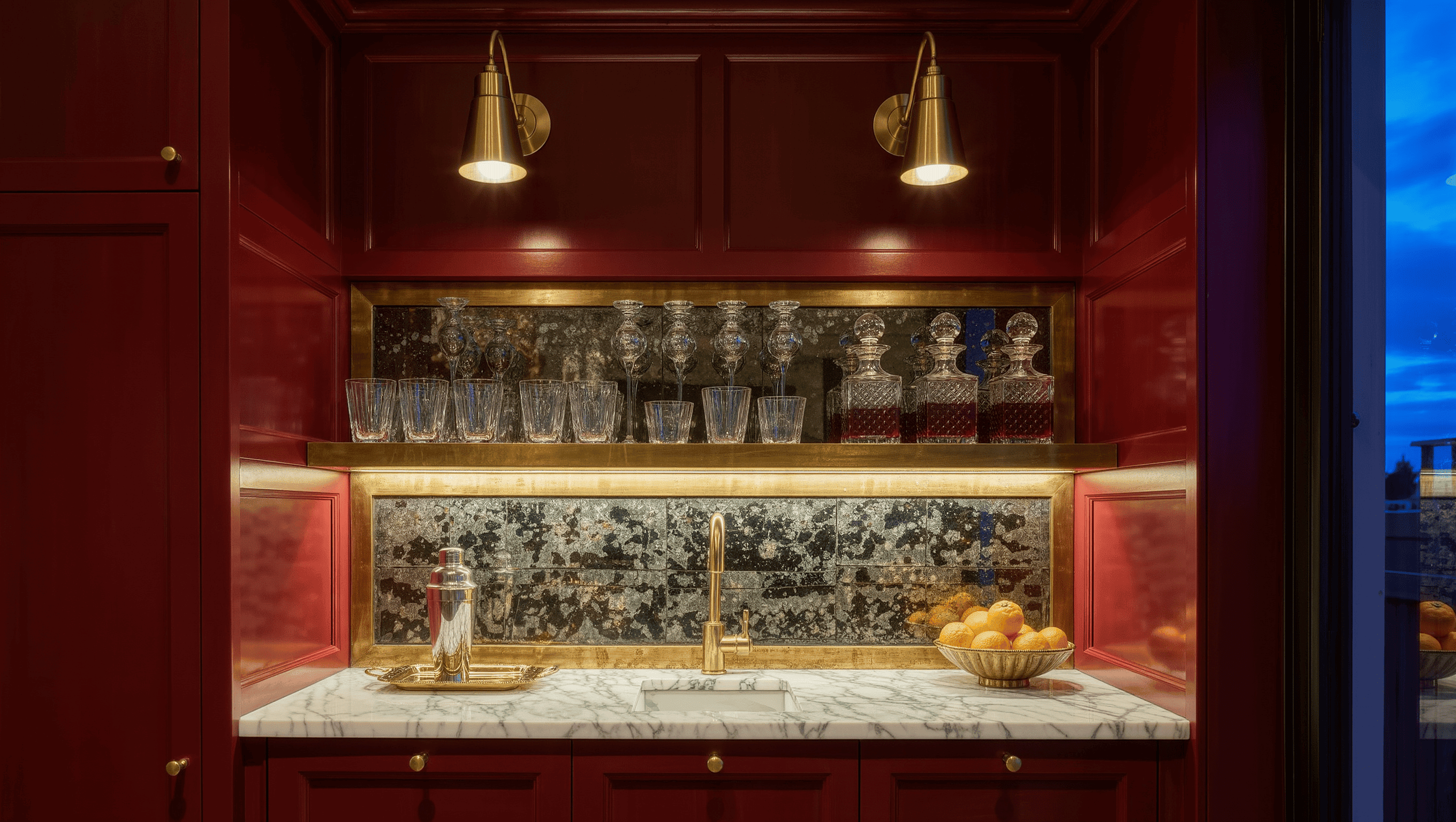 A home bar at dusk with an oxblood back wall, antique mirror panel, brass shelves of glassware, and a white marble counter.