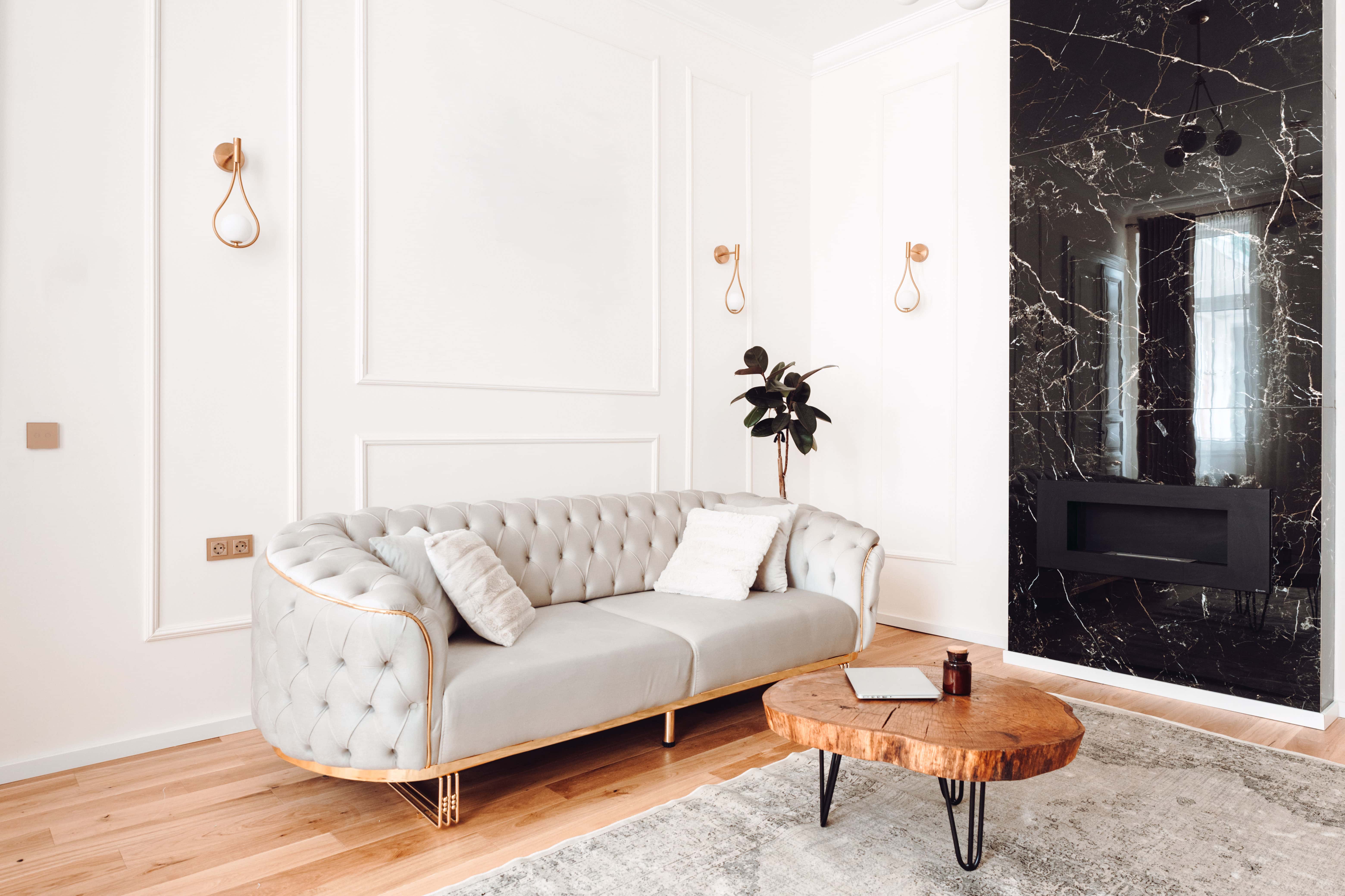 A tufted cream sofa sits beneath twin gold wall sconces, beside a round marble-topped side table and a dramatic black-and-gold marble wall panel.