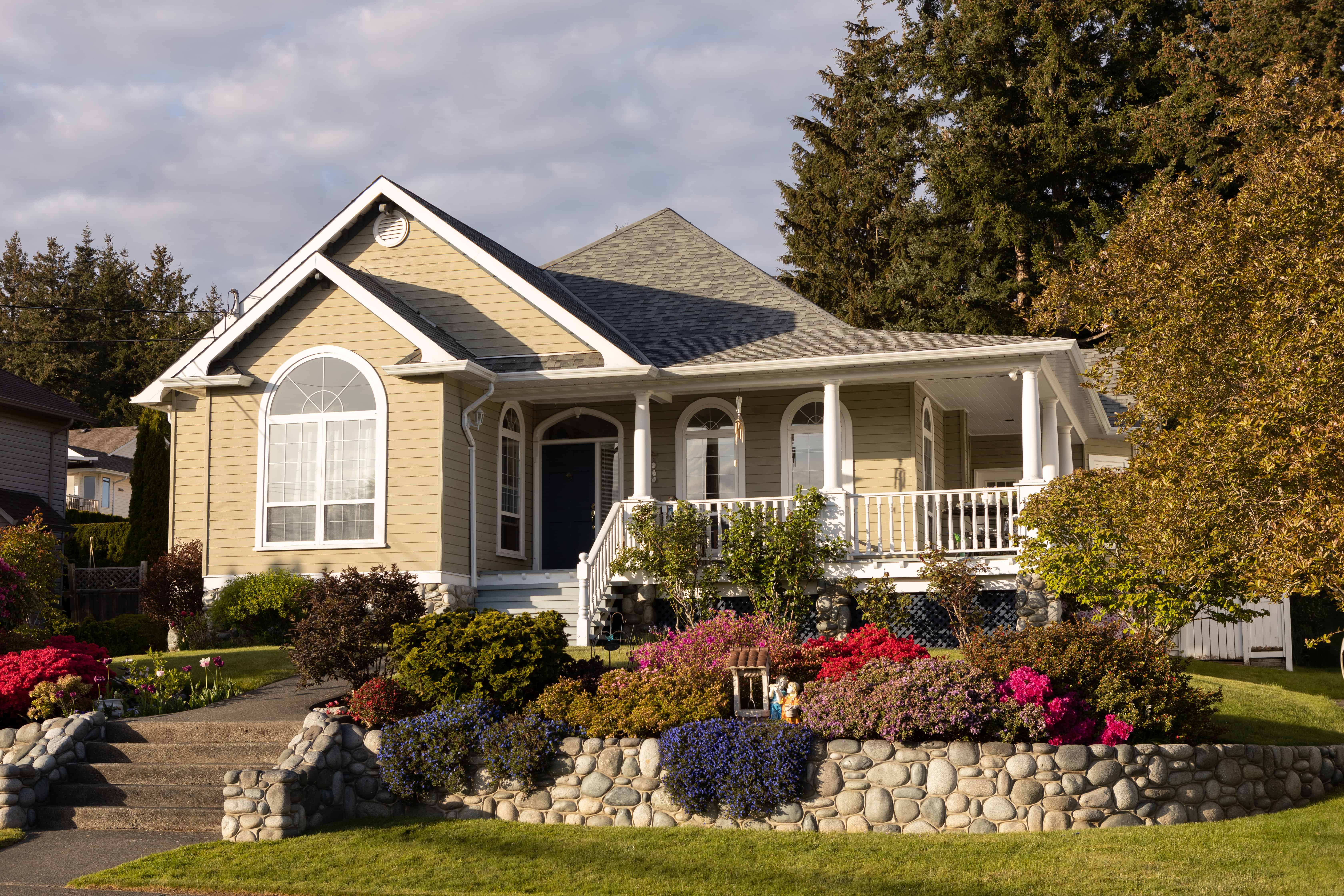 A stone retaining wall holds layered pink and red flowering shrubs that step down from the house's siding to the lawn.