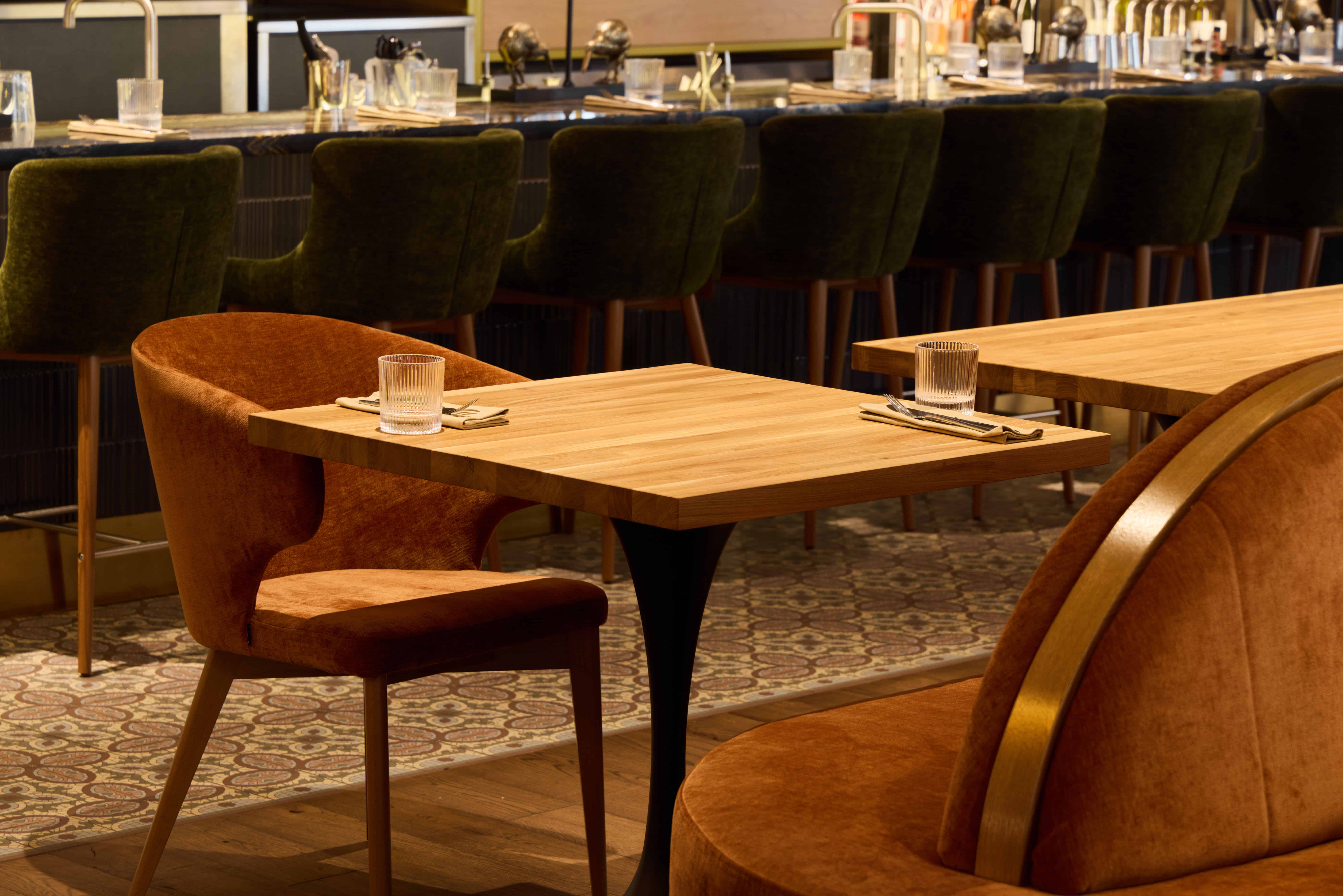Emerald velvet banquette seating lines a wooden counter, with mustard leather chairs and wood tables set for dining in warm ambient light.