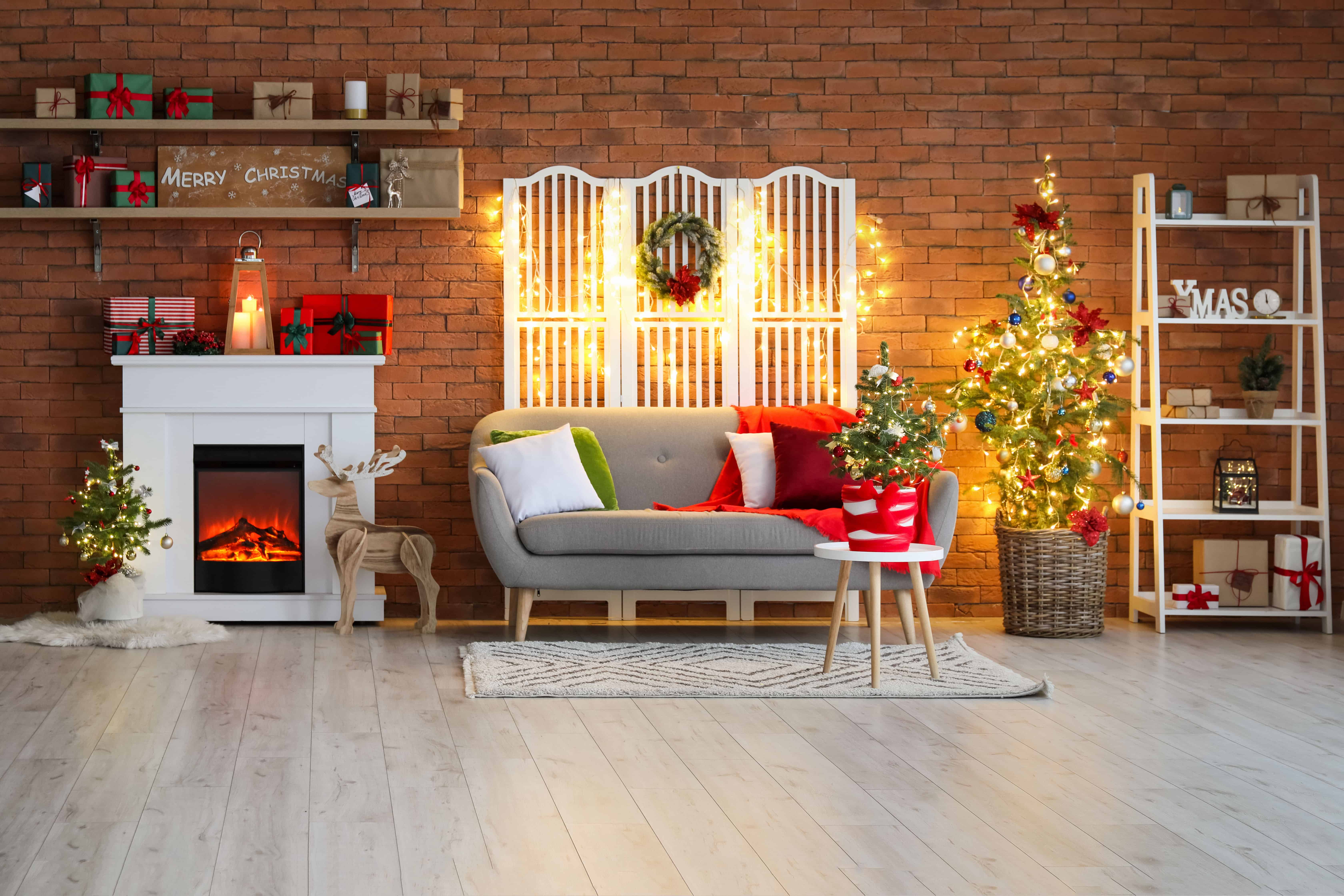 An open shelving unit beside a decorated tree holds mixed ornaments, wrapped gifts, and warm string lights against an exposed brick wall.