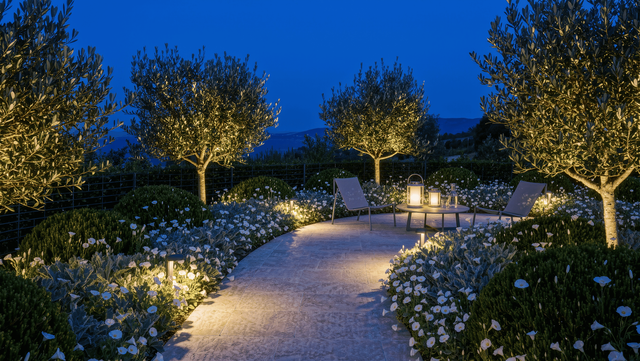 A dusk garden with a pale gravel path between white blooms and dark yew silhouettes, lit by low path lights and two enclosed storm lanterns on a table.