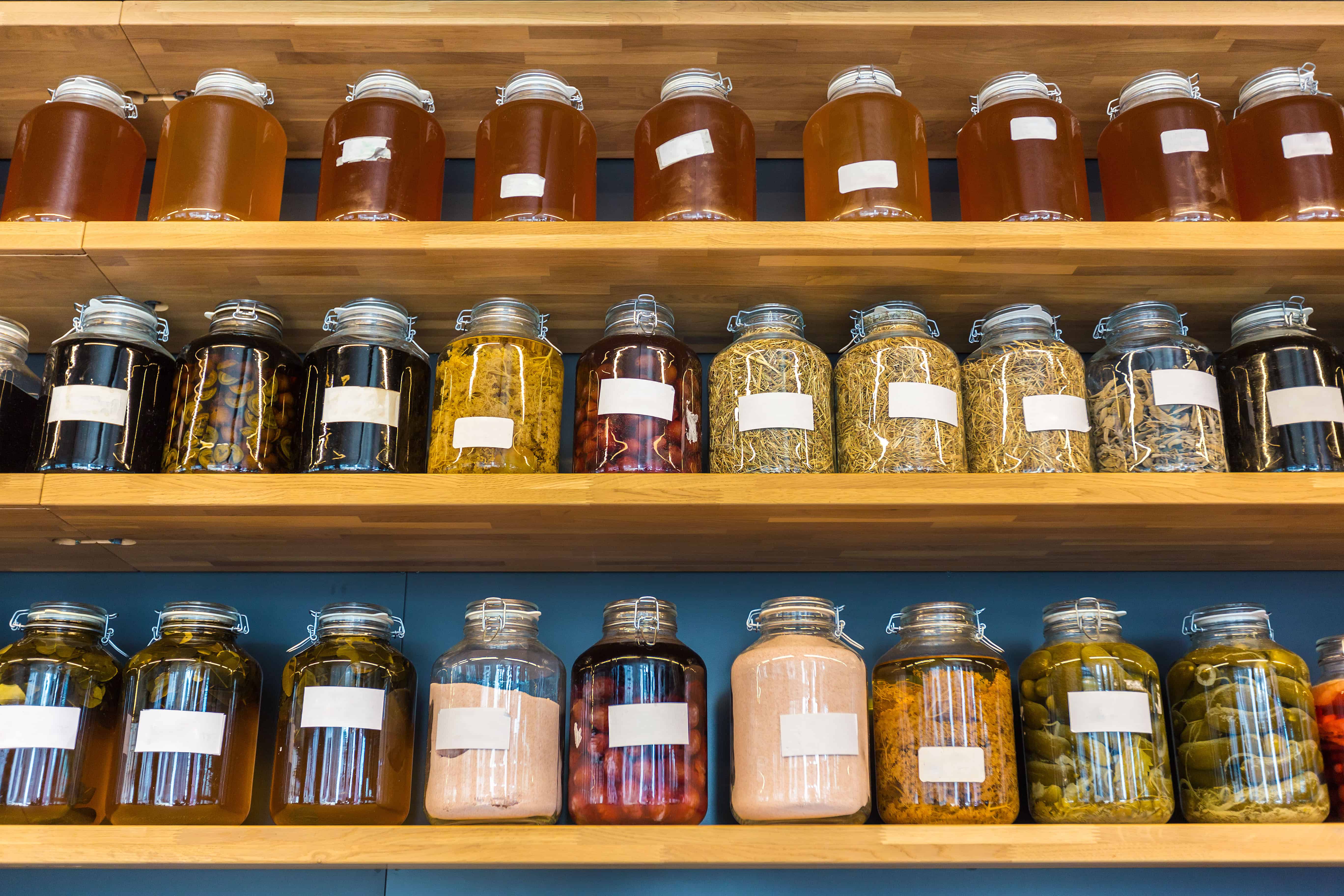 Rows of glass jars filled with preserves, grains, and pantry staples line wood shelves against a deep teal wall.