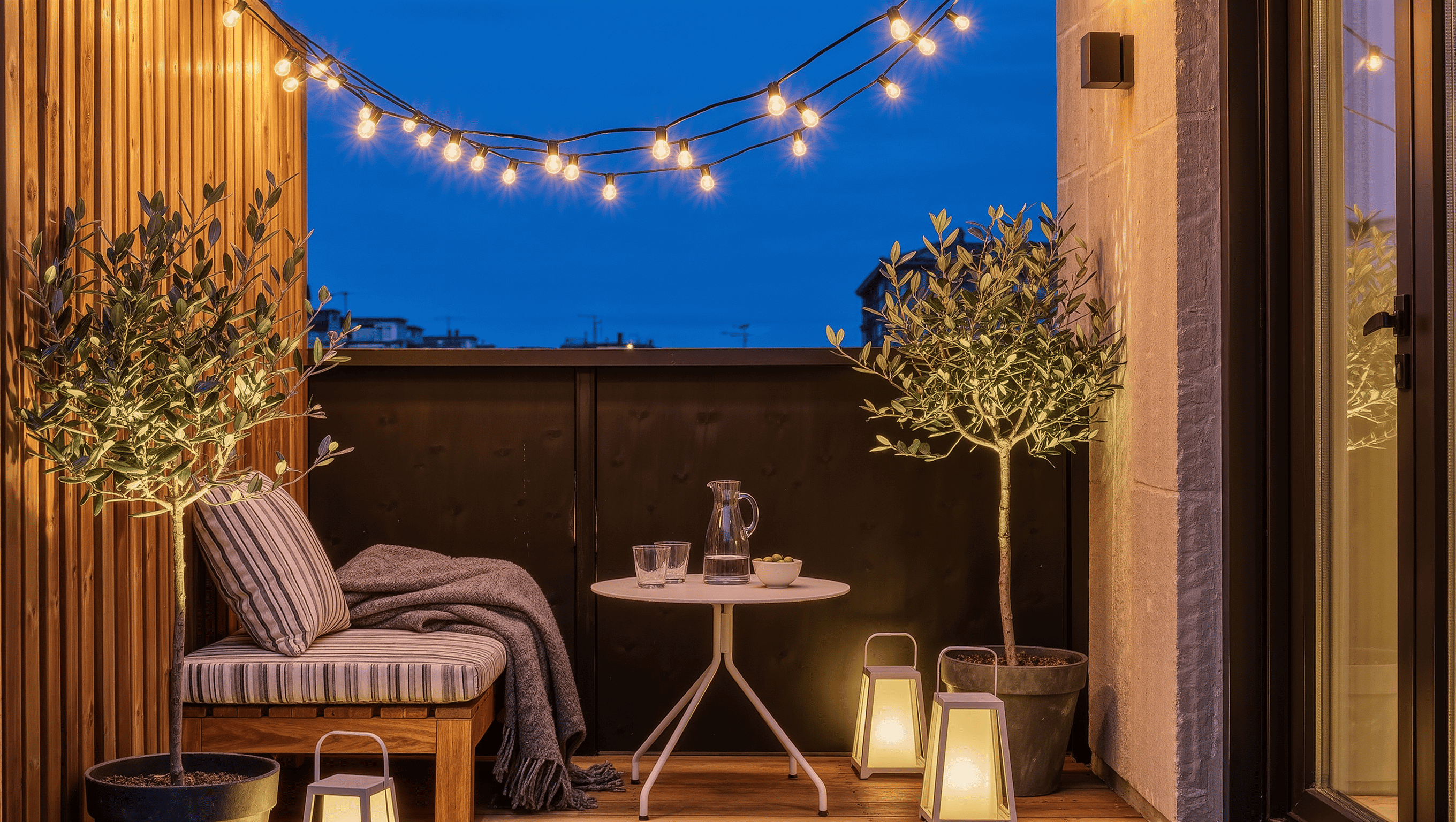 An urban patio at dusk with string lights overhead, enclosed storm lanterns by a cushioned bench, a bistro table, and potted olive trees.