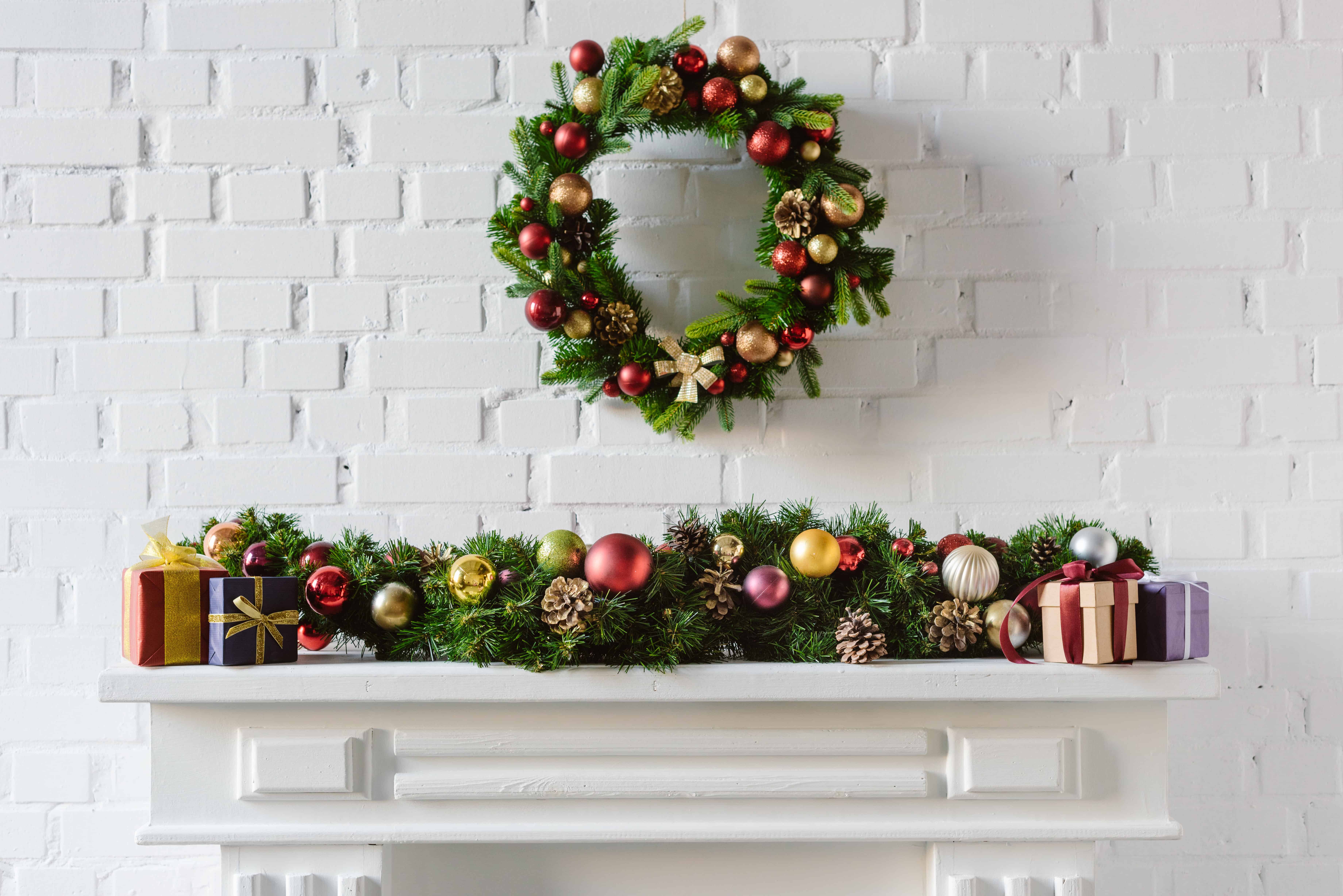 A white mantel is layered with a garland of baubles, wrapped gifts, and a matching wreath in red, gold, and green above it.