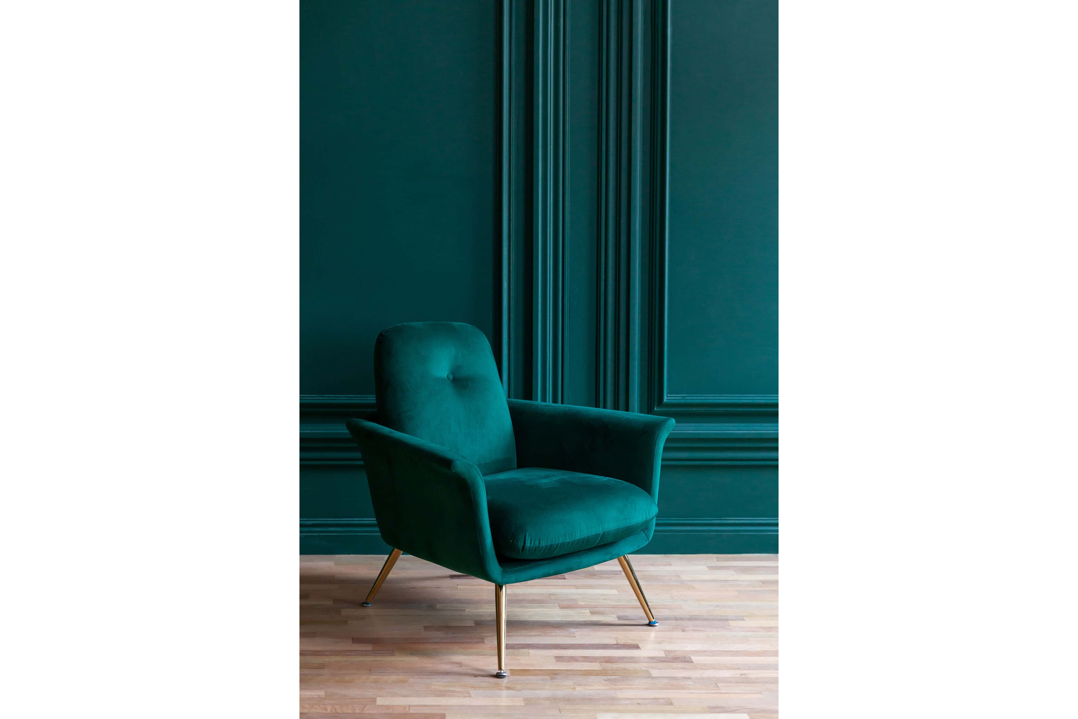 A tufted emerald green velvet armchair with brass legs sits against a matching emerald green paneled wall.