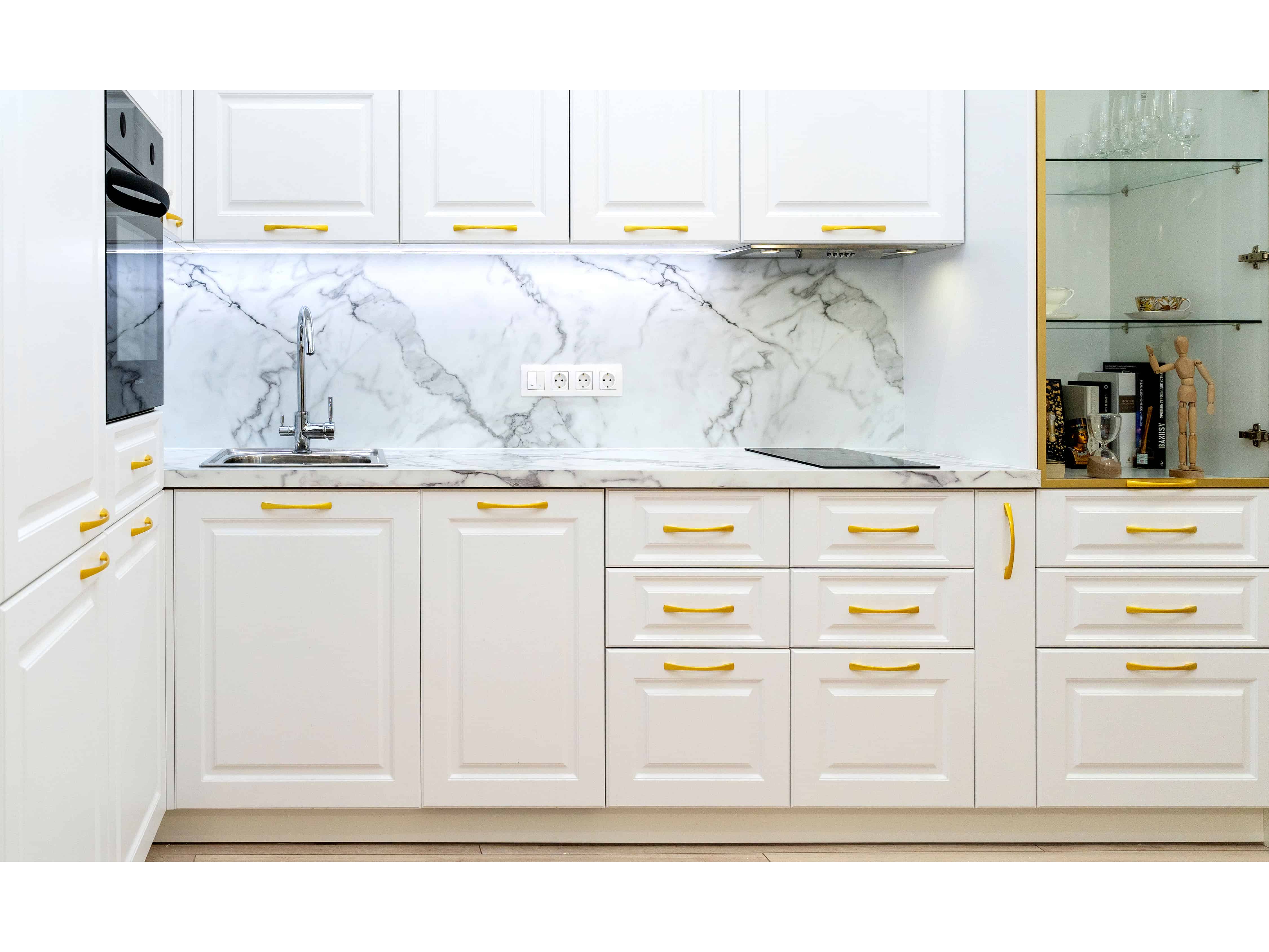 A row of white shaker cabinets with brass hardware surrounds a marble backsplash, keeping cookware and dishware tucked behind matching doors.