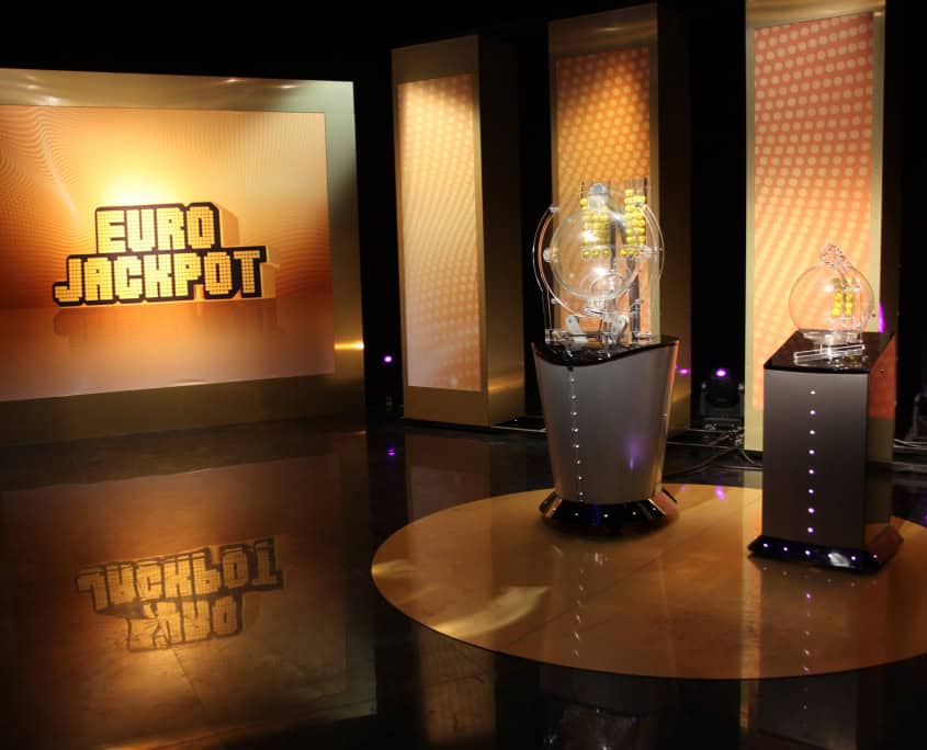 Eurojackpot TV studio with illuminated draw machines and a glowing logo on a golden backdrop before a lottery drawing.