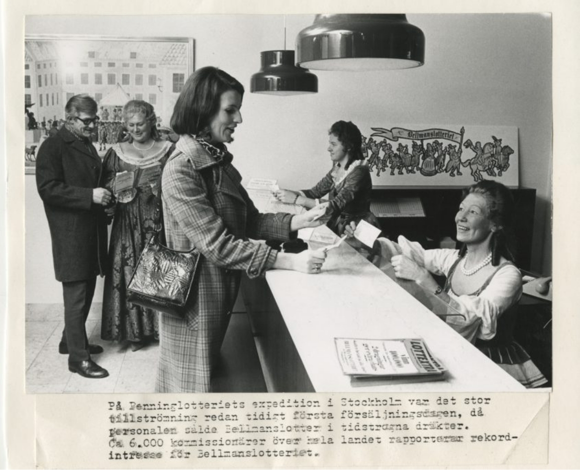 People in period costumes and contemporary clothing stand at a counter in a Stockholm office, receiving lottery tickets from staff dressed in historical attire, with signage for the Bellmanlotteriet lottery visible in the background. The scene is from the first sales day of the Bellmanlotteriet, a historic Swedish lottery.