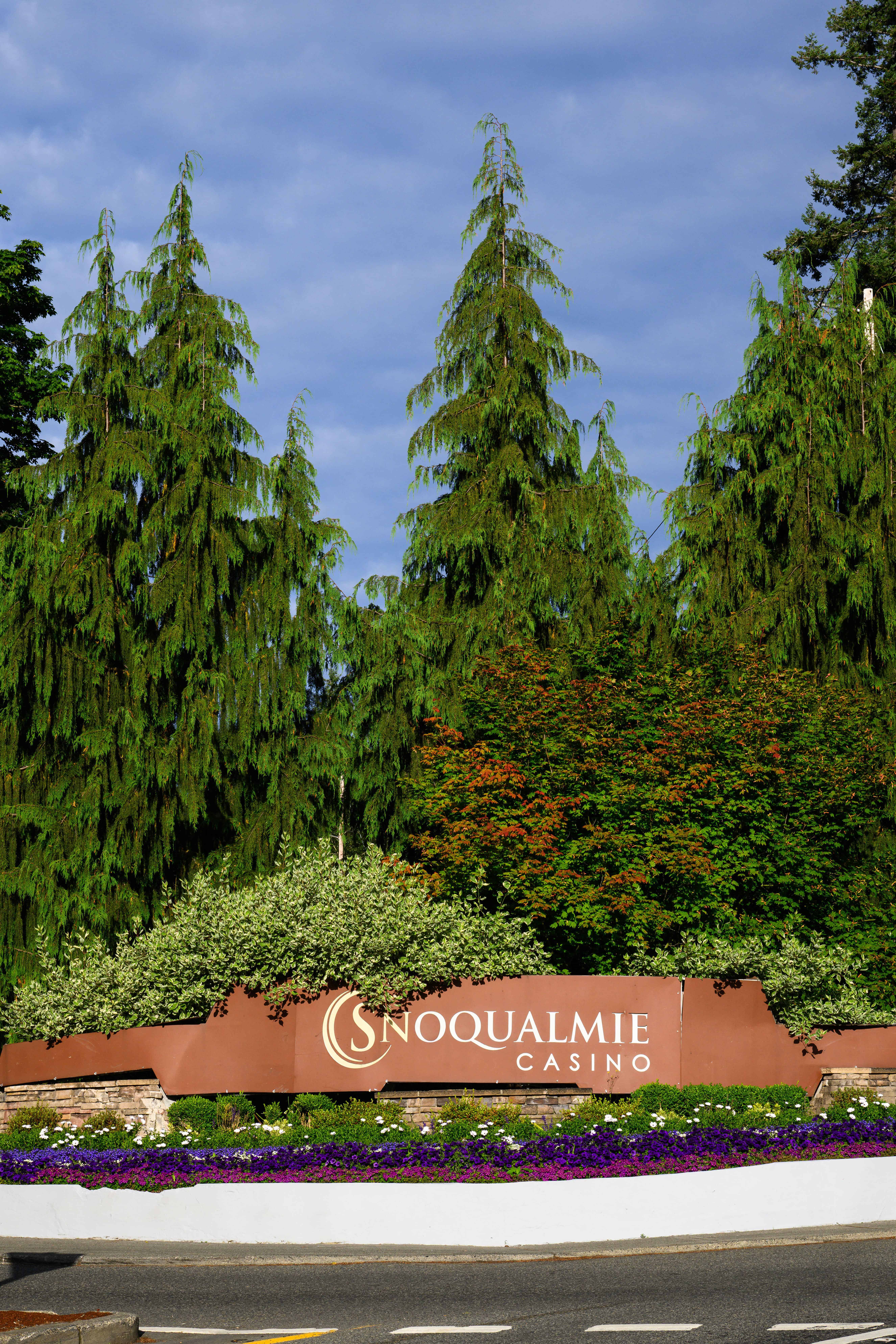Entrance sign for Snoqualmie Casino in Washington State, surrounded by tall Pacific Northwest conifers and flowering gardens against a blue sky