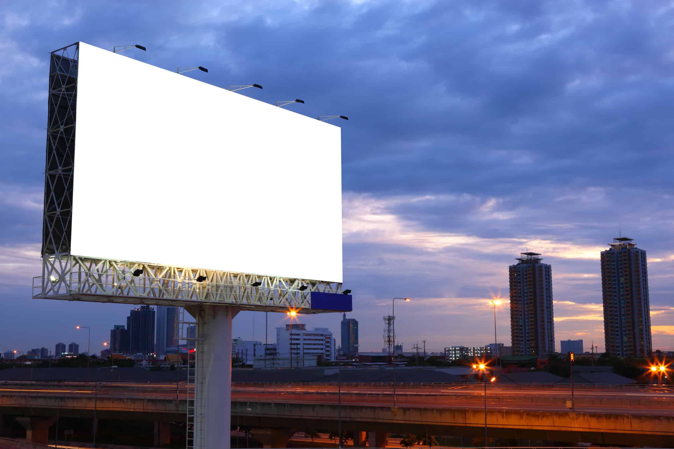 An empty roadside billboard against a city skyline at dusk.