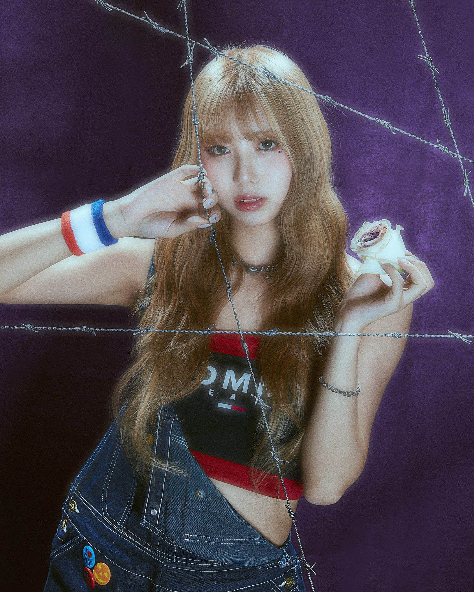 An Asian woman with long dark blonde hair, in a red, white and blue tube top and denim overalls holds up a white rose behind barbed wire.