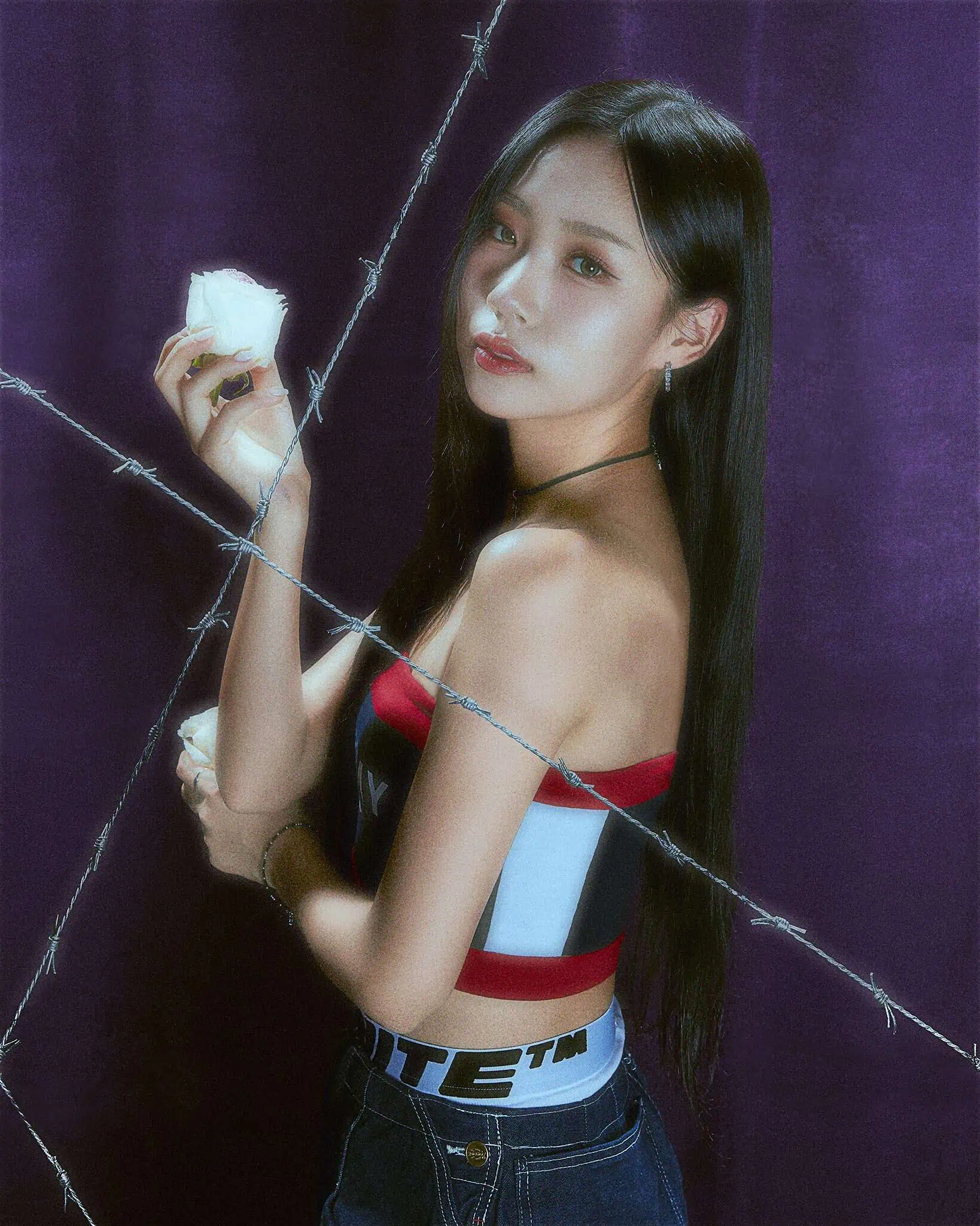 An Asian woman with black hair in a red, white and blue tube top and jeans holds up a white rose behind barbed wire.