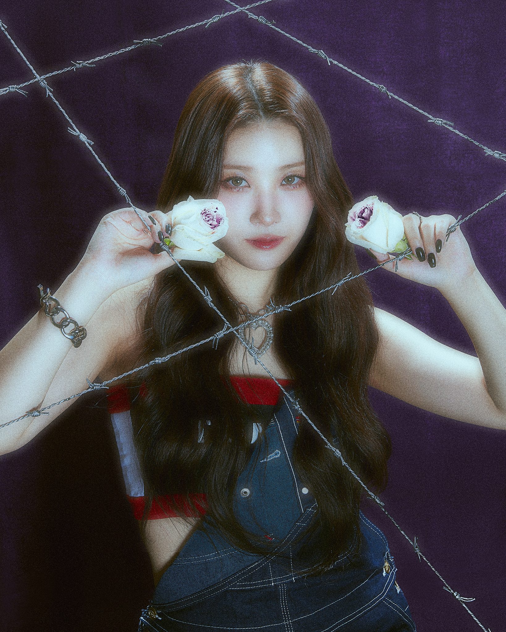 An Asian woman with brown hair in a red, white and blue tube top and denim overalls holds up white roses behind barbed wire.