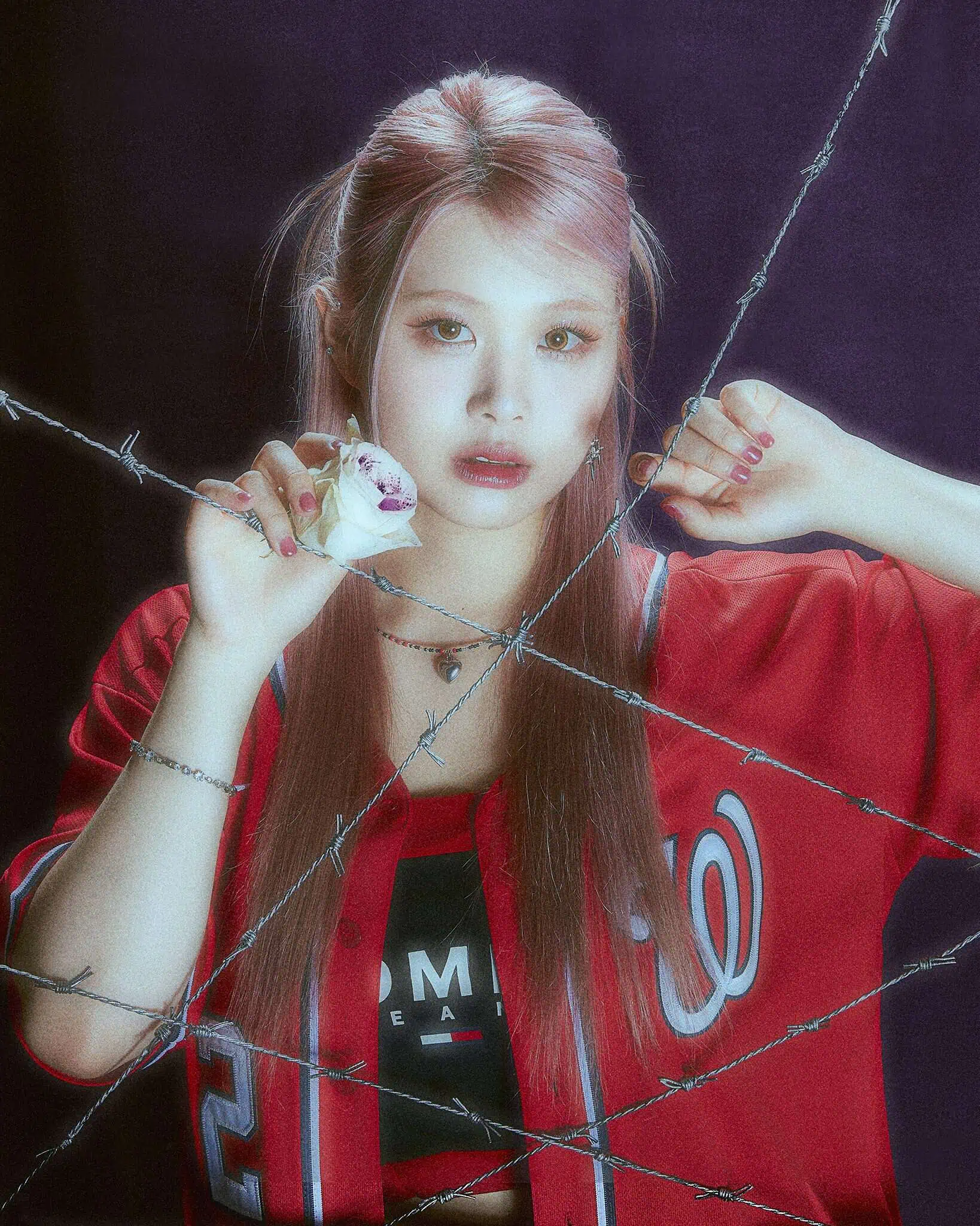 An Asian woman with light reddish brown hair in a half ponytail and a red baseball jersey holds up a white rose behind barbed wire.