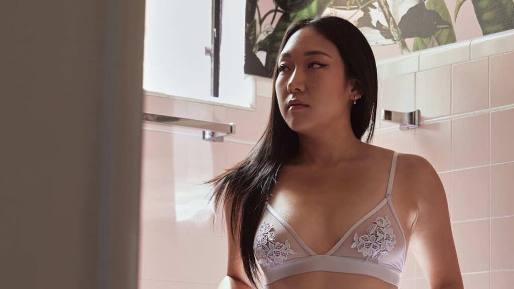 A closeup of an Asian American woman wearing a sheer, pale purple bra, in a pink-tiled bathroom.