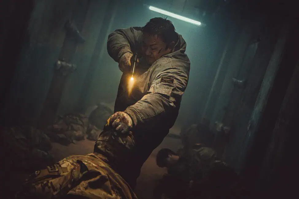 Ma Dong-seok as Nam San in "Badland Hunters" holds a sparking gun to the head of a person dressed in camouflage, with other persons laying down in the background.