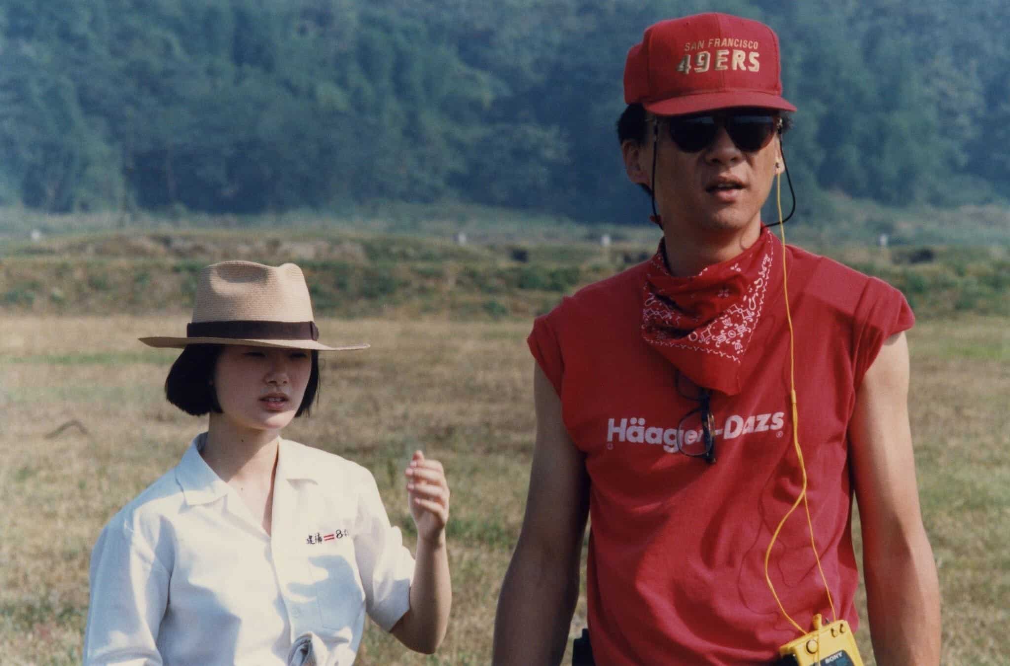 An Asian woman in a white top and beige brimmed hat and an Asian man in a red t-shirt with "Haagen-Dazs" and red San Francisco 49ers hat stand in a field with greenery in the background.