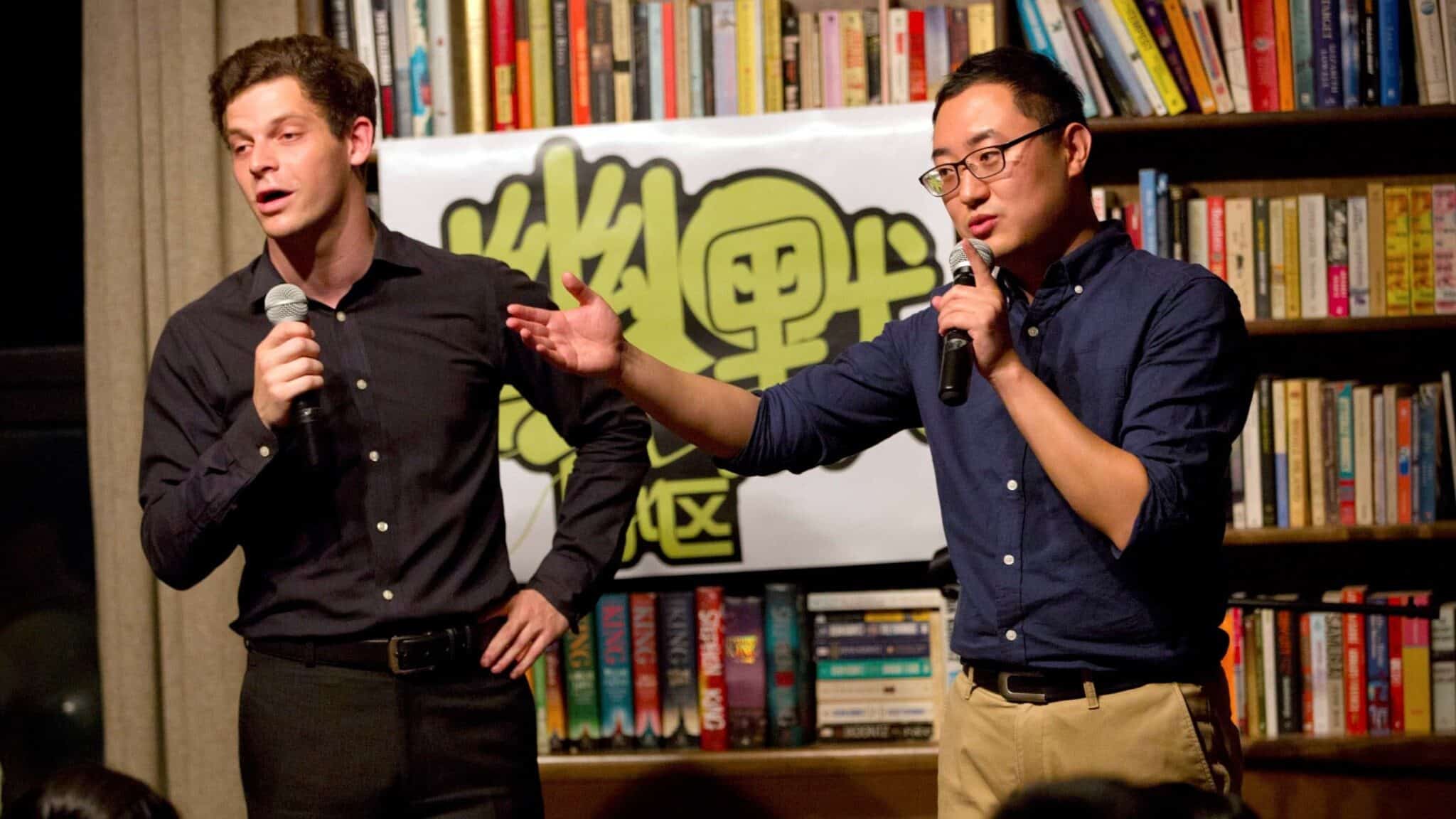 A white man in black clothing and an Asian man in a blue shirt and khaki pants, hold microphones to their mouths, with a bookshelf in the background.