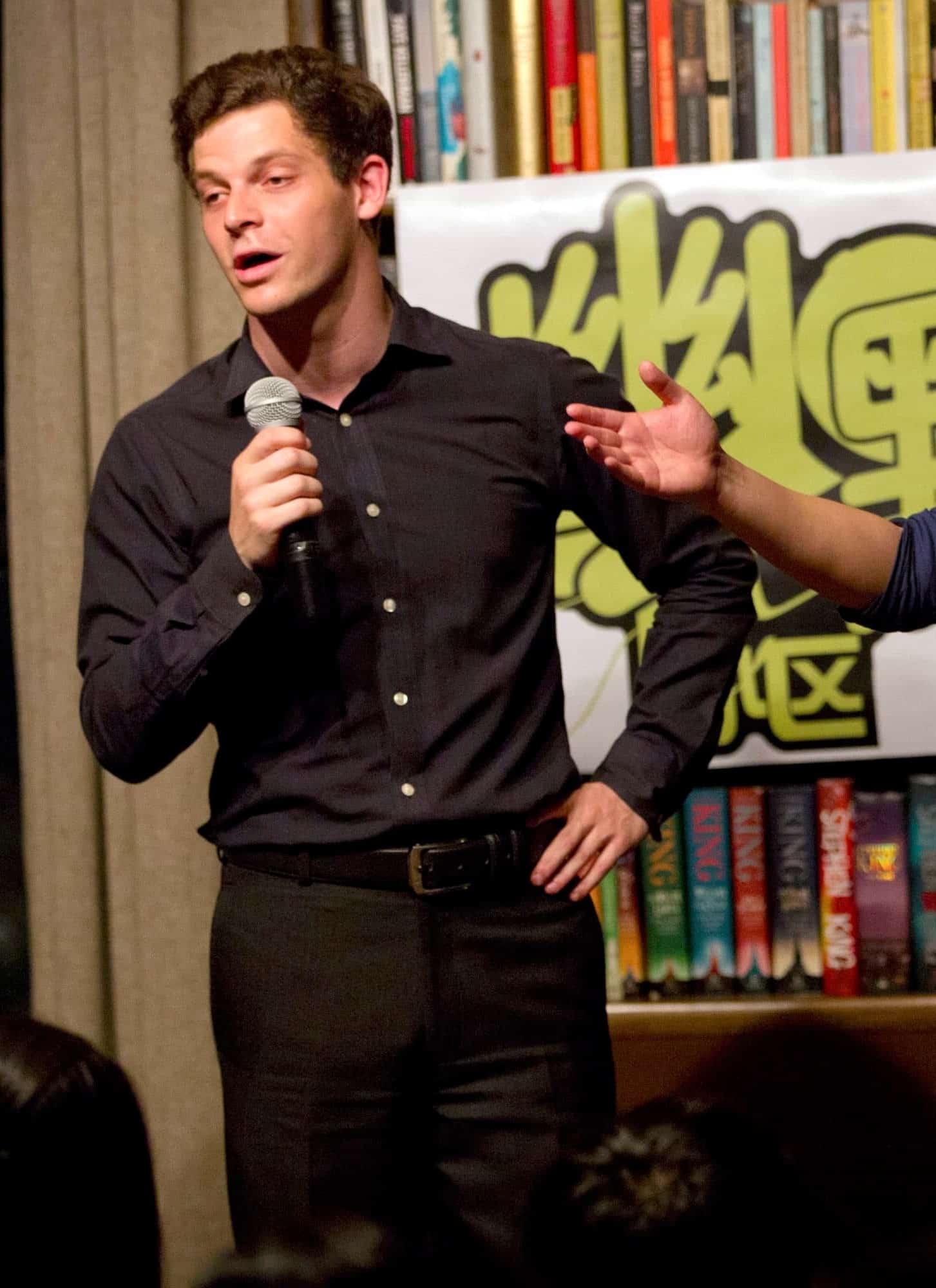 A white man in black clothing and an Asian man in a blue shirt and khaki pants, hold microphones to their mouths, with a bookshelf in the background.