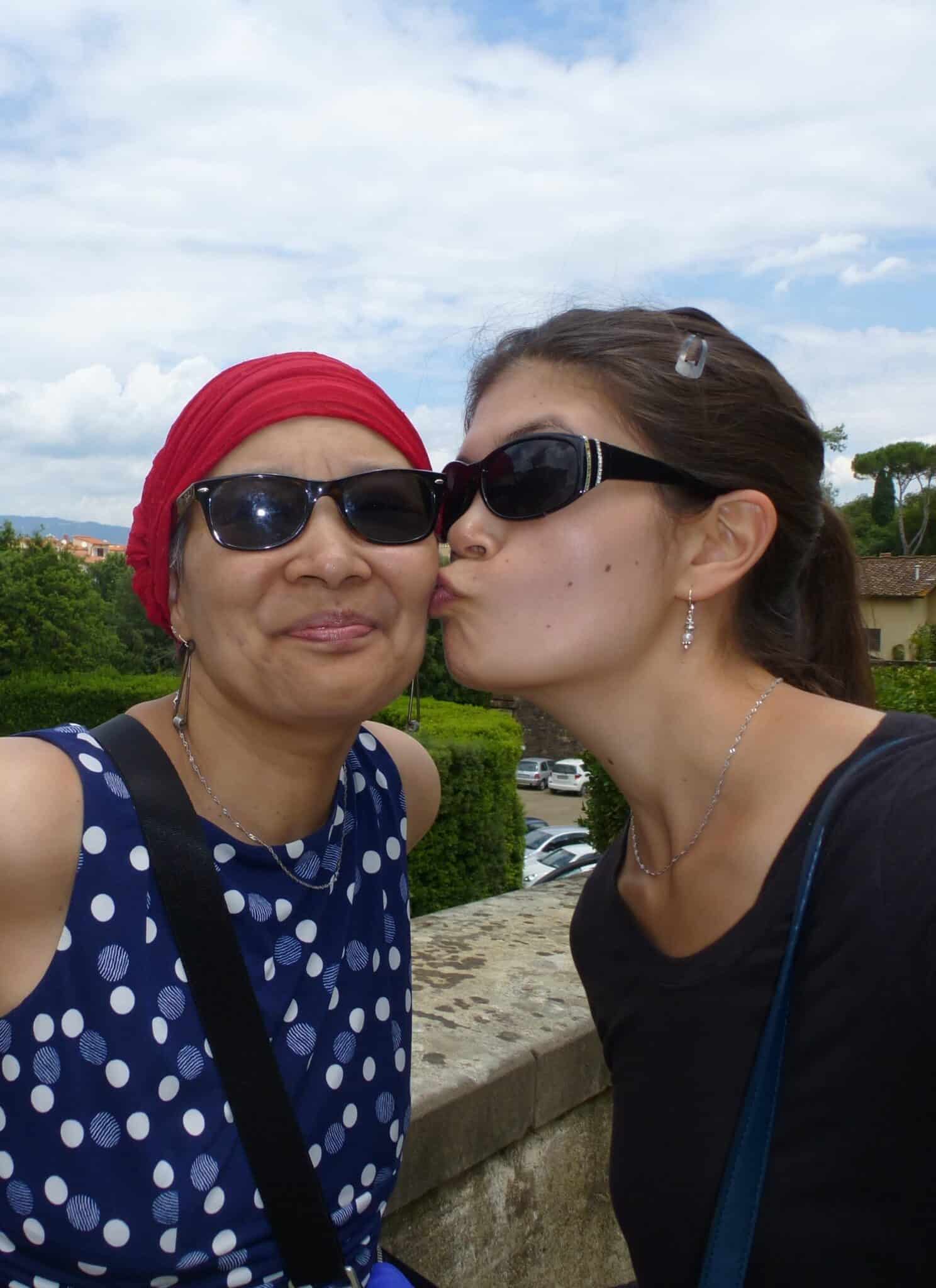 An Asian woman in a black top and sunglasses kisses an older Asian woman in a blue top, red head wrap and sunglasses on the cheek, with houses and a blue sky in the background.