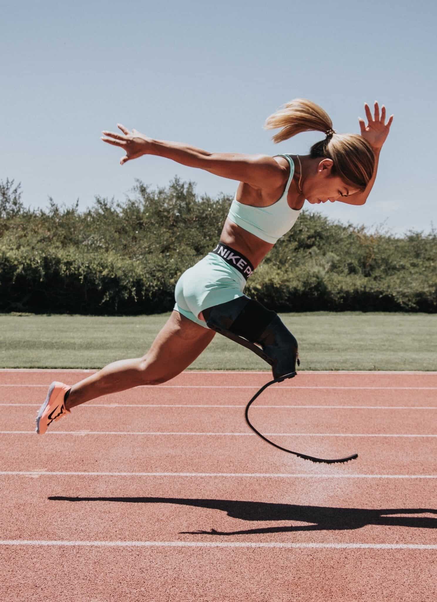 Para athlete Scout Bassett, in a light blue top and shorts, runs on a track with a black running blade on her right side.