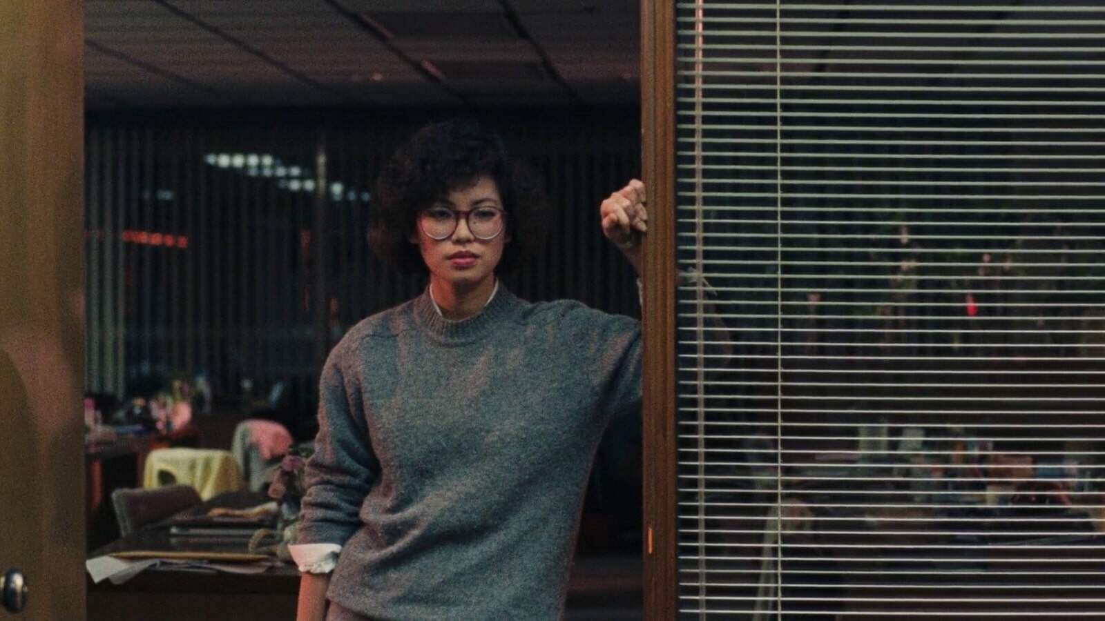 An Asian woman with short curly hair, with glasses and a gray sweater leans against a doorframe in an office.