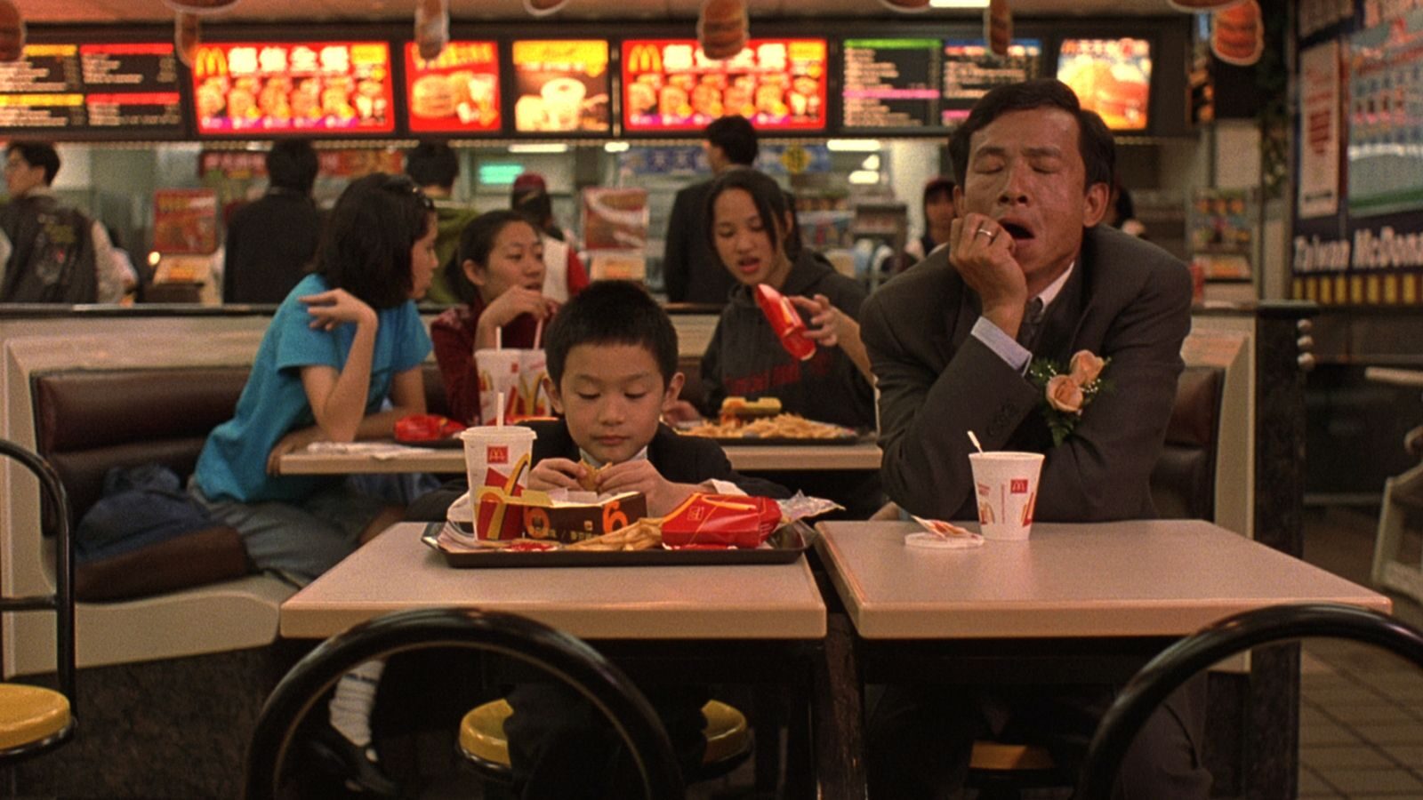 An Asian boy and man sit at two tables in McDonald's in suits, with food in front of the boy and the man is asleep.