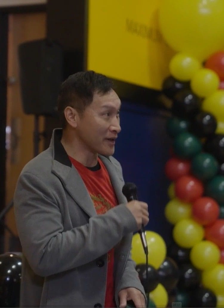 A Black woman and Asian man stand with a microphone in their hands with balloons in the background.