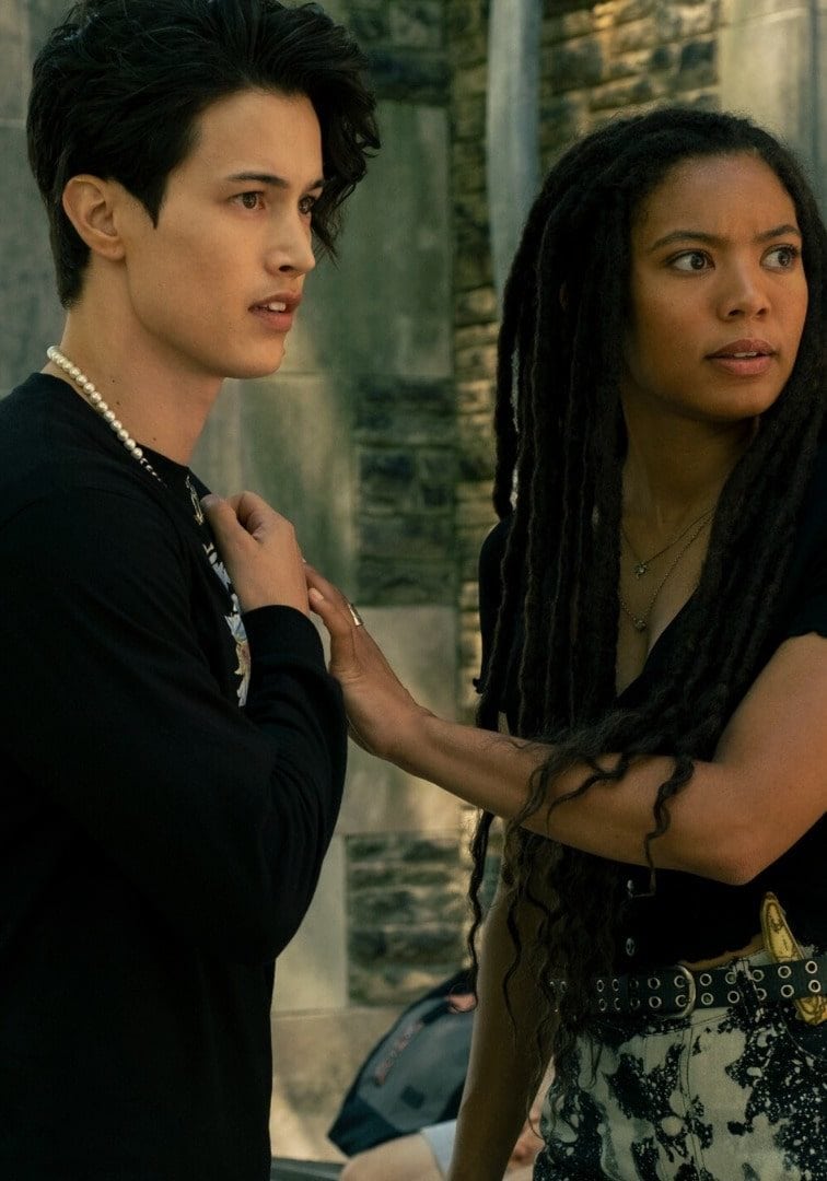 A young woman and a young masculine individual stand outside a building. The woman is grabbing the front of their shirt. Both look startled by something off camera.
