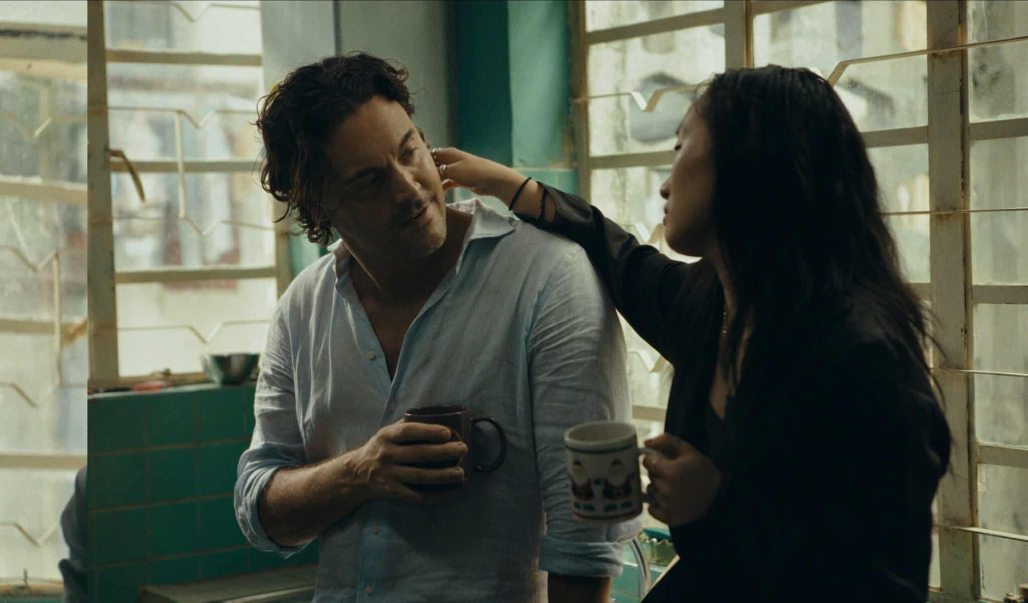 A woman sits together with a man in a kitchen, each holding a mug. The woman has her hand reached out to the man's face in an intimate, relaxed gesture.
