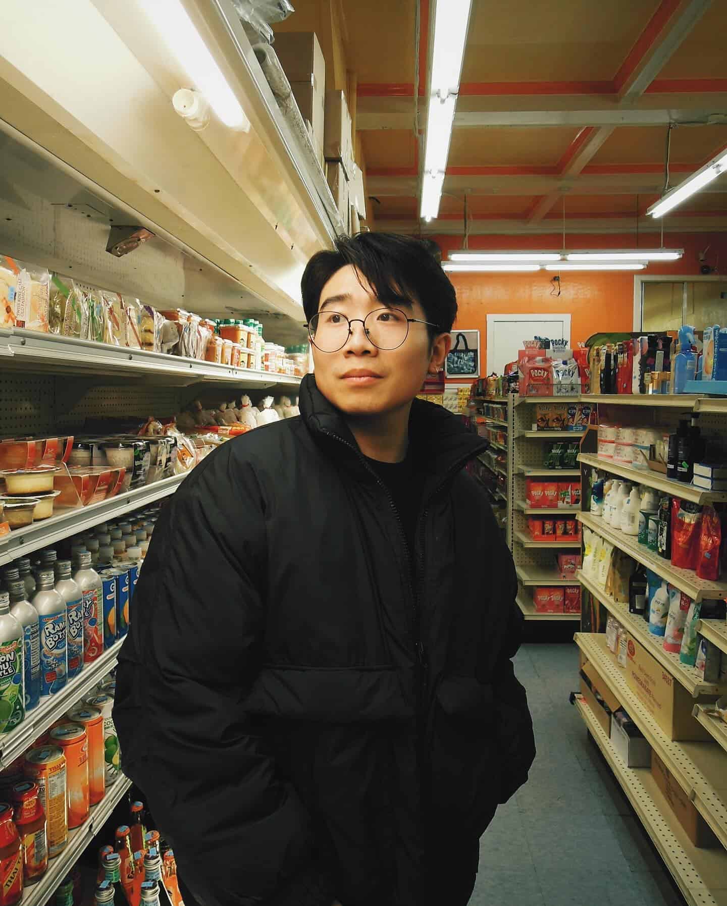 A man wearing a winter coat stands in a grocery store aisle, looking pensively into the distance.