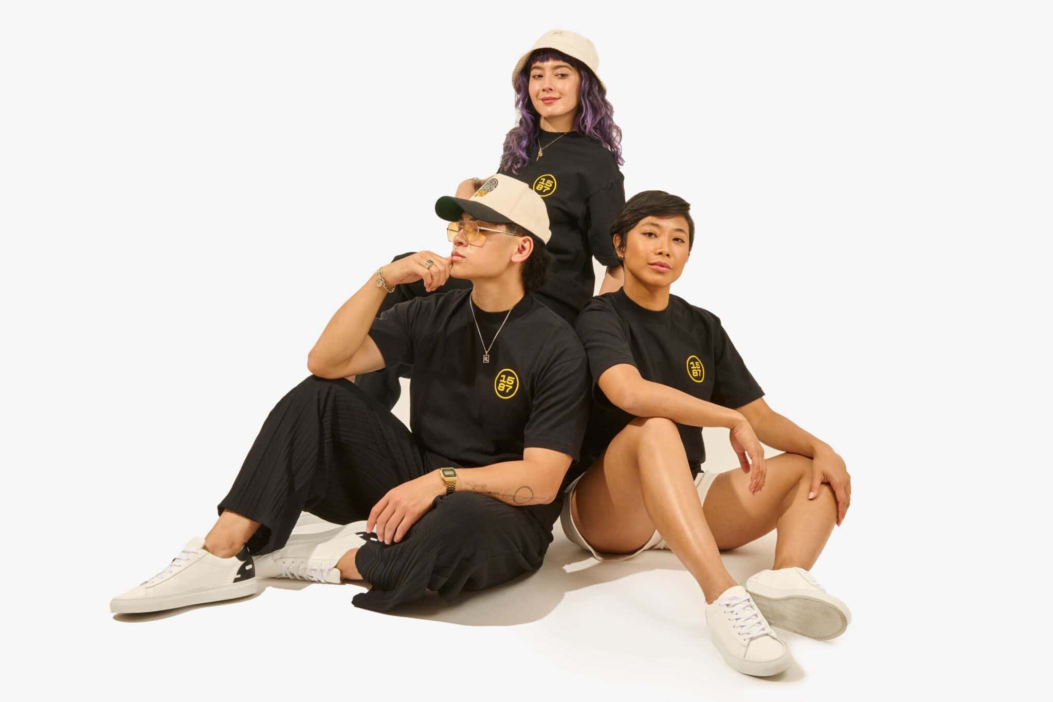 Three Asian people sit and stand together in black outfits, with "1587" on their t-shirts, against a white background.
