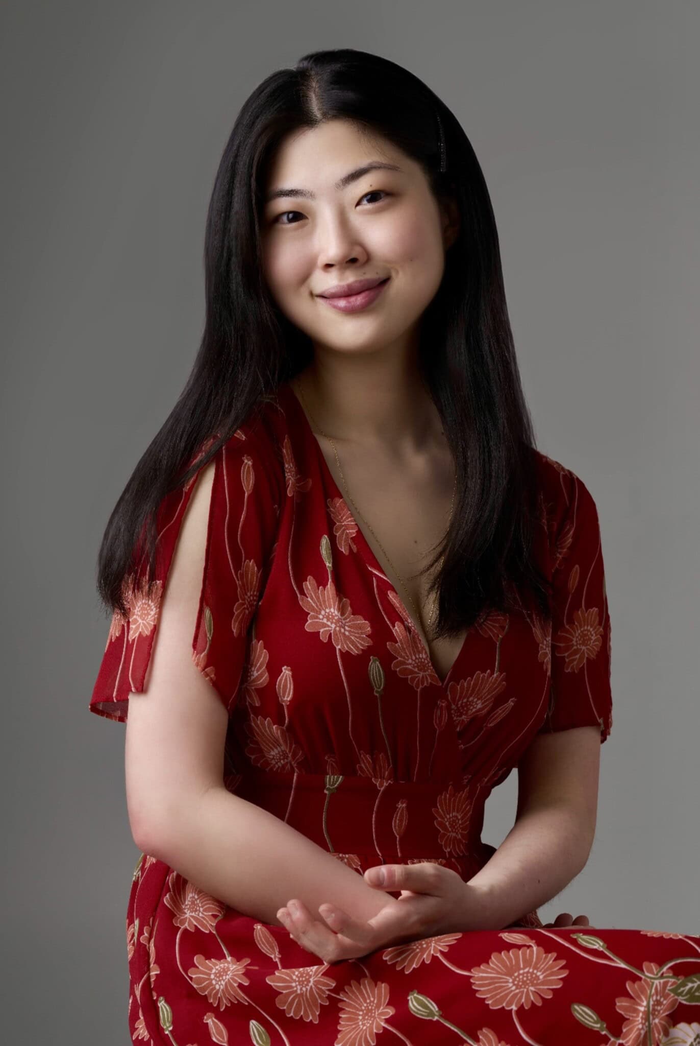 A portrait of author Kyla Zhao, with long black hair, in a red floral dress, against a gray background.