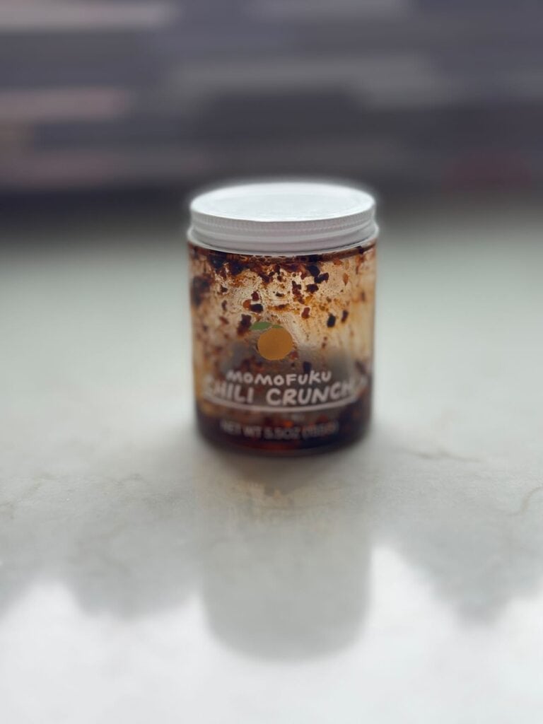 Momofuku Chili Crunch in an almost empty jar on a white tabletop.