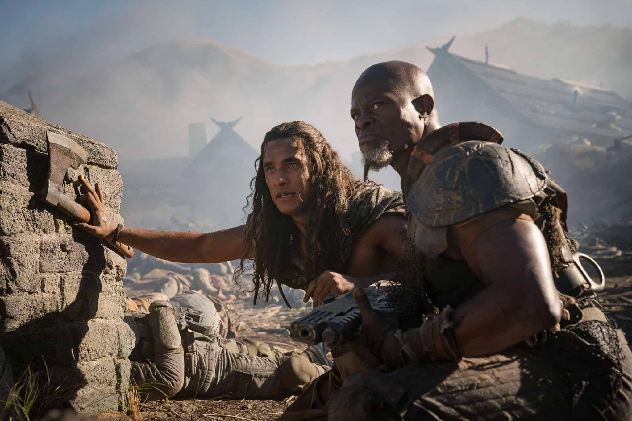 Actors Staz Nair and Djimon Hounsou in "Rebel Moon — Part Two: The Scargiver," in a battlefield with smoke and houses in the background.