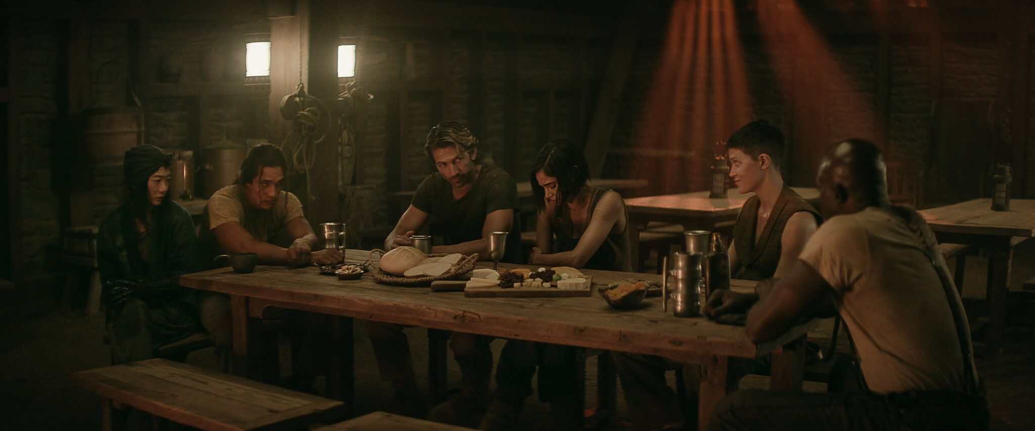 A scene from "Rebel Moon — Part Two: The Scargiver," with a group of people sitting at a long table with food and drinks on top.