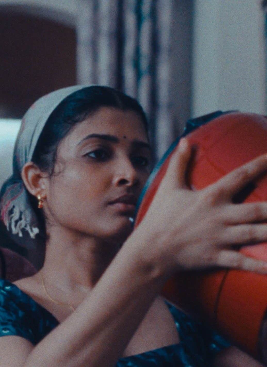 A south Asian woman in a dark green top, with a light gray scarf around her hair, holds up a red-orange rice cooker.