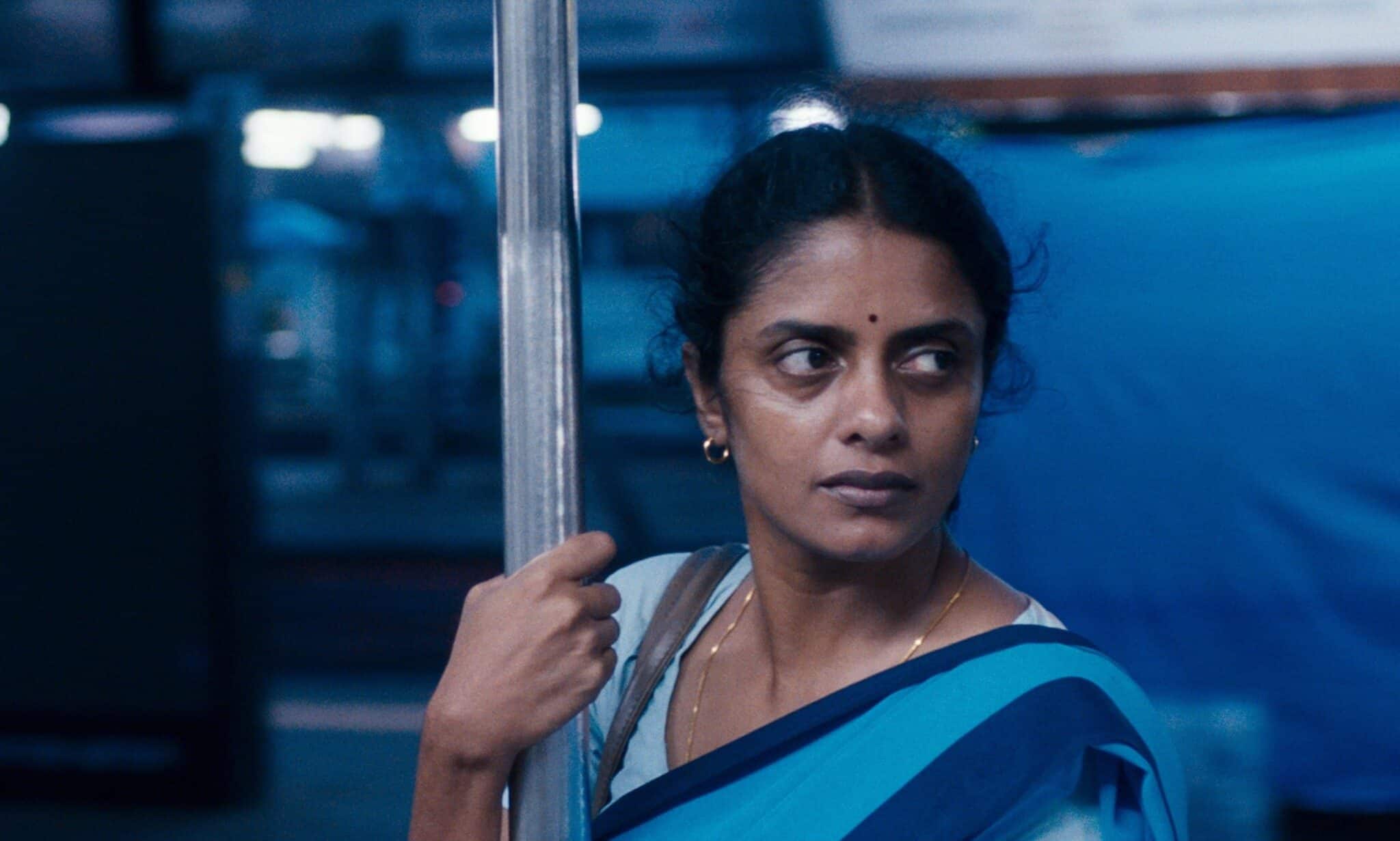 A south Asian woman in blue, stands holding a silver pole, against a blue backdrop.